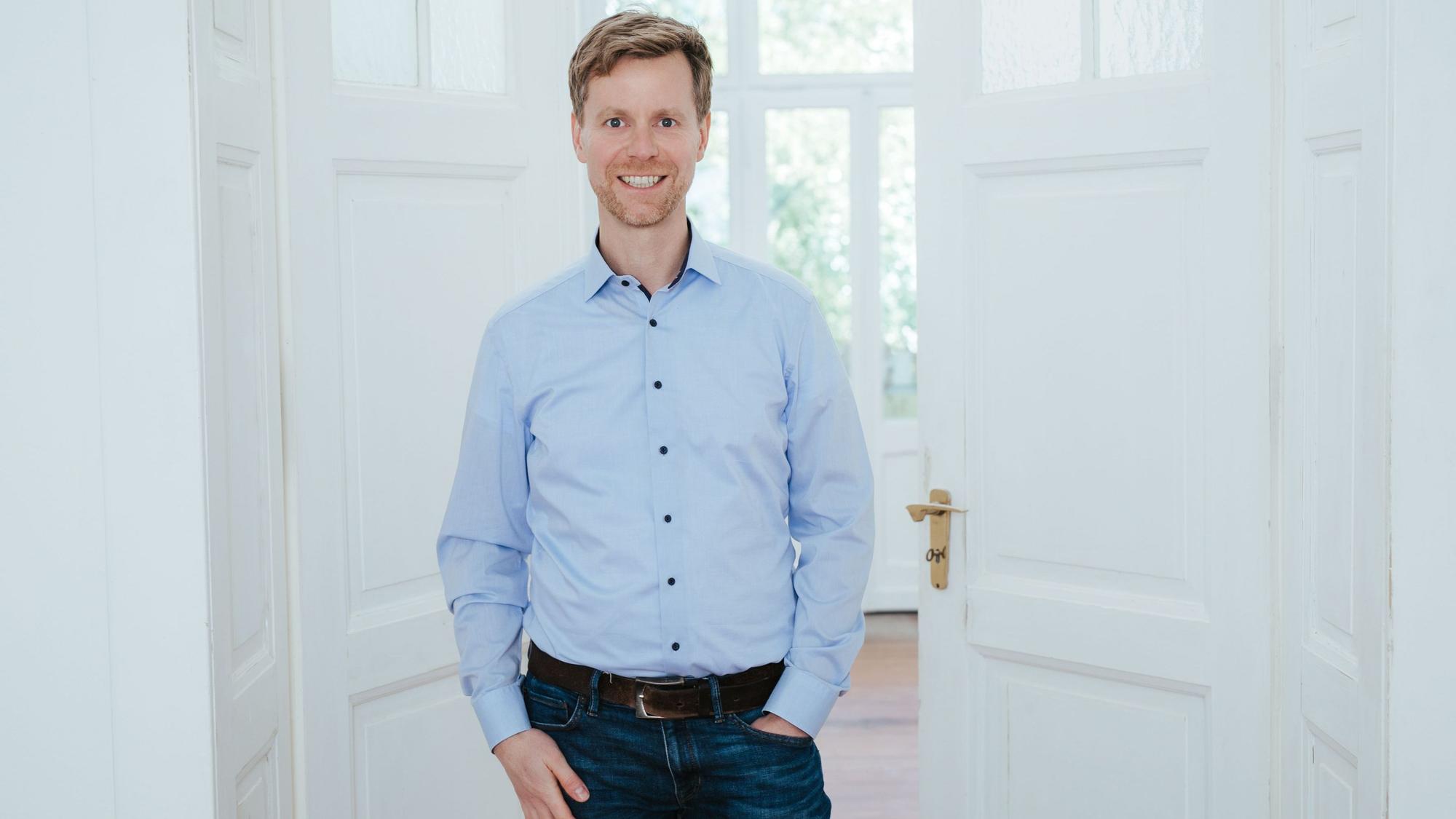Ein Mann in blauem Hemd vor einer weißen, halb geöffneten Tür.