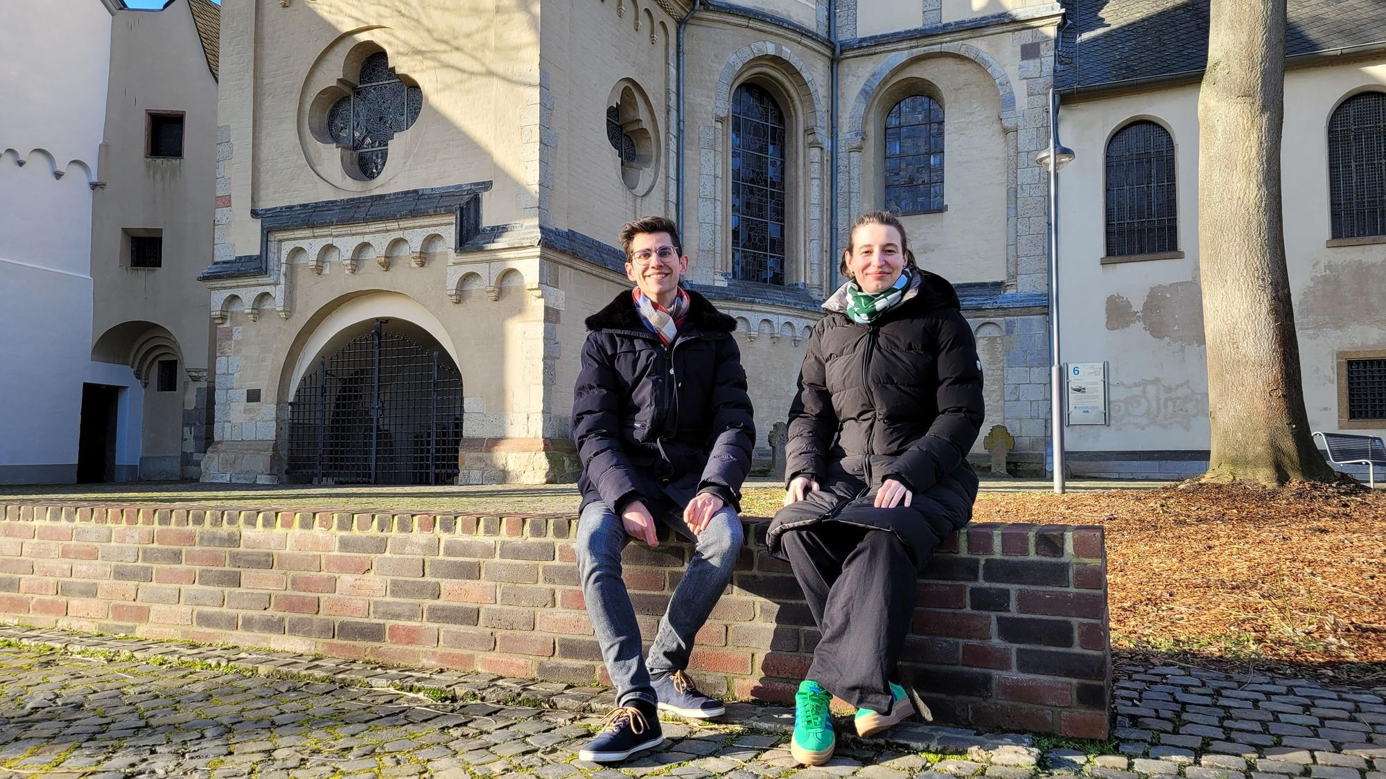 Ein Frau und ein Mann sitzen auf einer Mauer. Im Hintergrund die Abteikirche St. Nikolaus in Brauweiler.