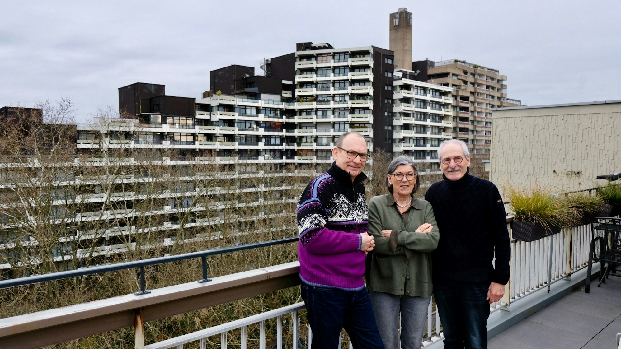 Zwei Männer und eine Frau stehen auf einem Balkon, im Hintergrund ist ein großer Wohnblock zu sehen.