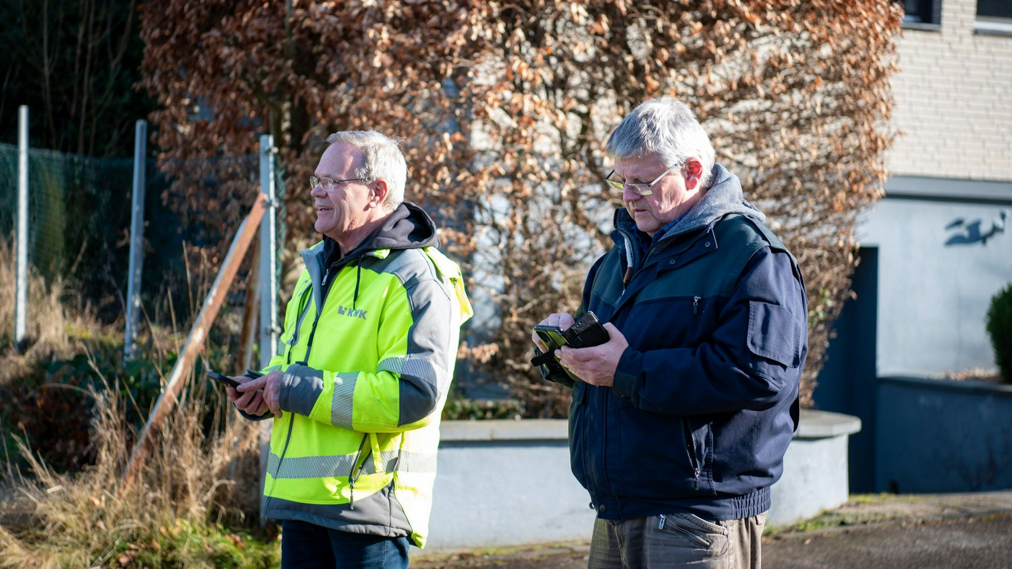 Zwei Männer stehen vor einer braunen Hecke nebeneinander. Beide haben Smartphones in der Hand.