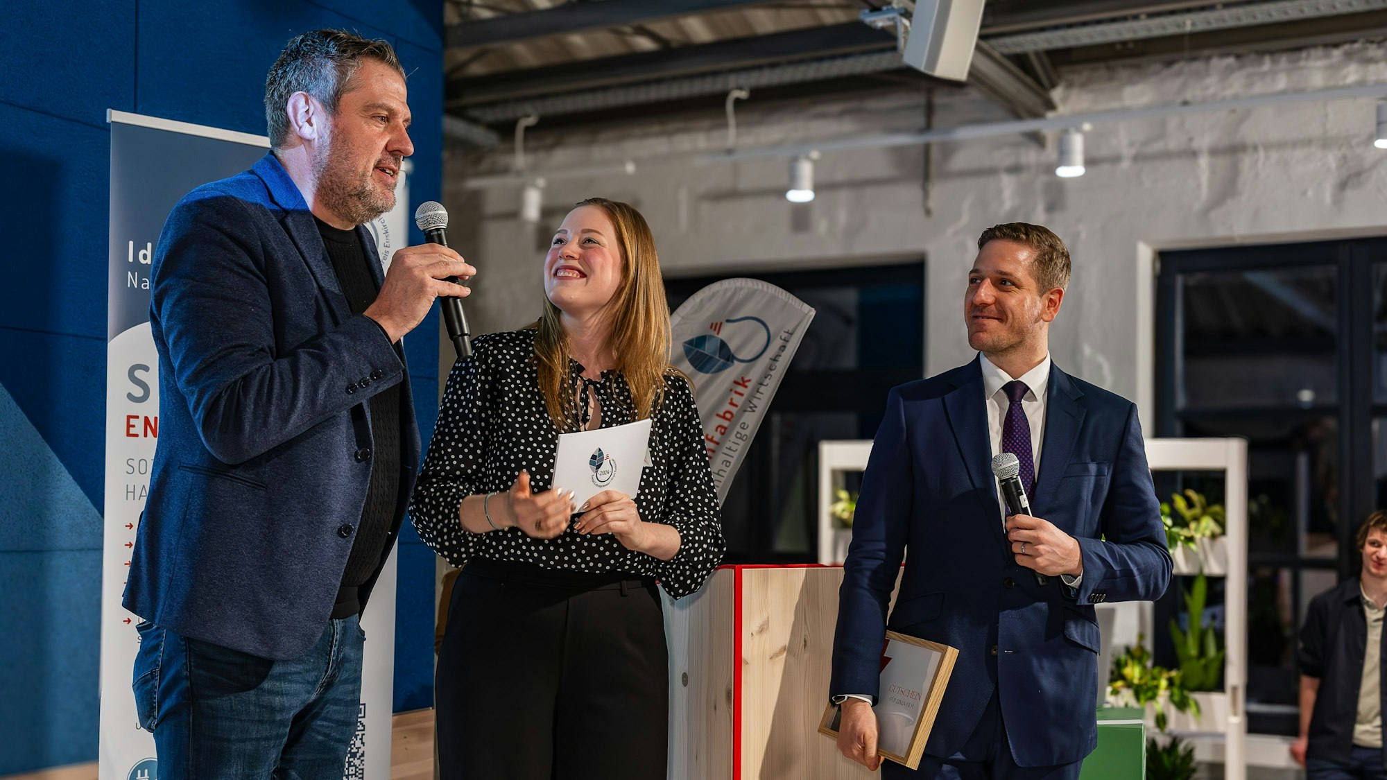 Lars Holocher bedankt sich am Mikro auf der Bühne für die Ehrung,die Moderatorin und der Landrat hören ihm zu.