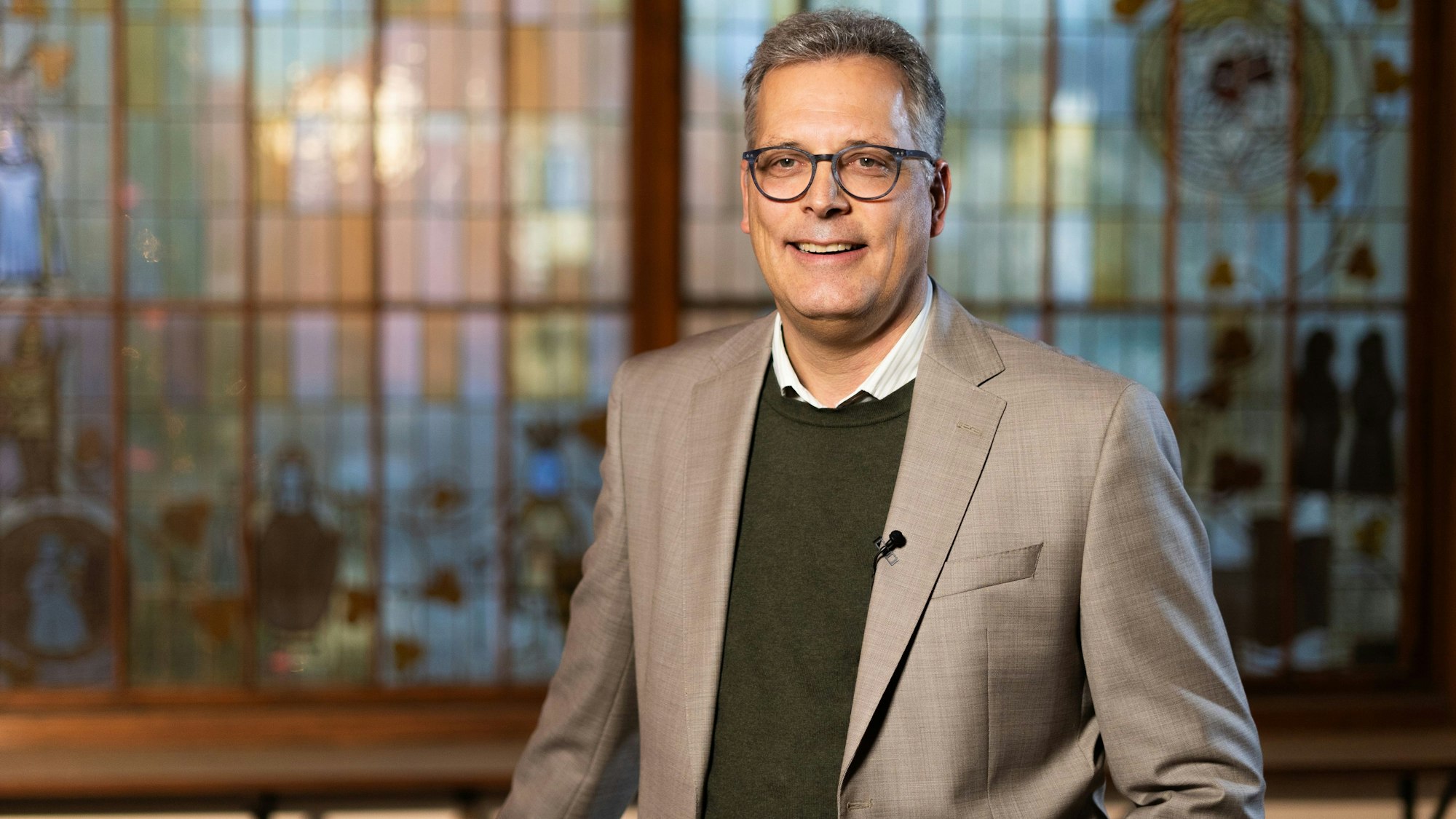 Bürgermeister Ulf Hürtgen (CDU) steht vor einem Buntglas-Fenster. Er trägt einen dunkelgrünen Pullover, darunter ein weißes Hemd und darüber ein graues Sakko.