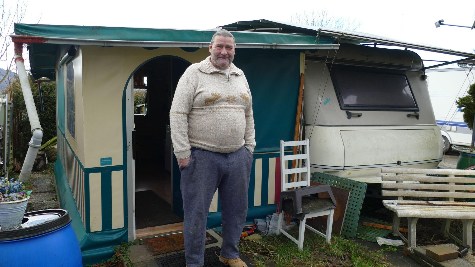 Ein Mann, der einen Pullover mit zwei Hirschen auf Brusthöhe trägt, steht vor einem kleinen Haus an einem Wohnwagen.