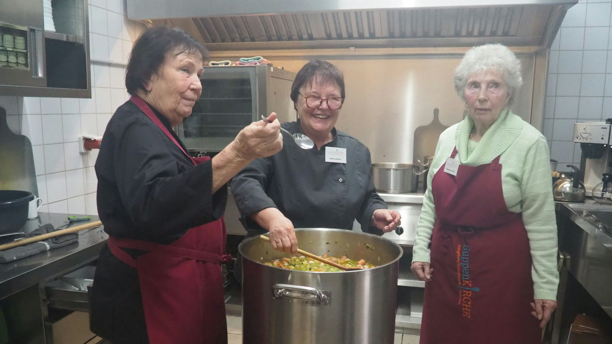 Die drei Köchinnen in der Küche, auf dem Herd ein großer Topf mit Suppe.