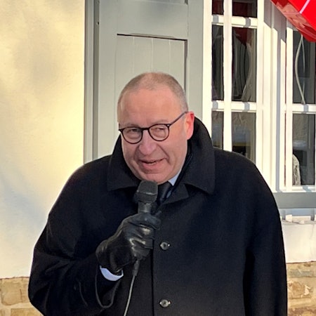 Ein Mann im schwarzen Mantel und Lederhandschuhen spricht in ein Mikrophon.