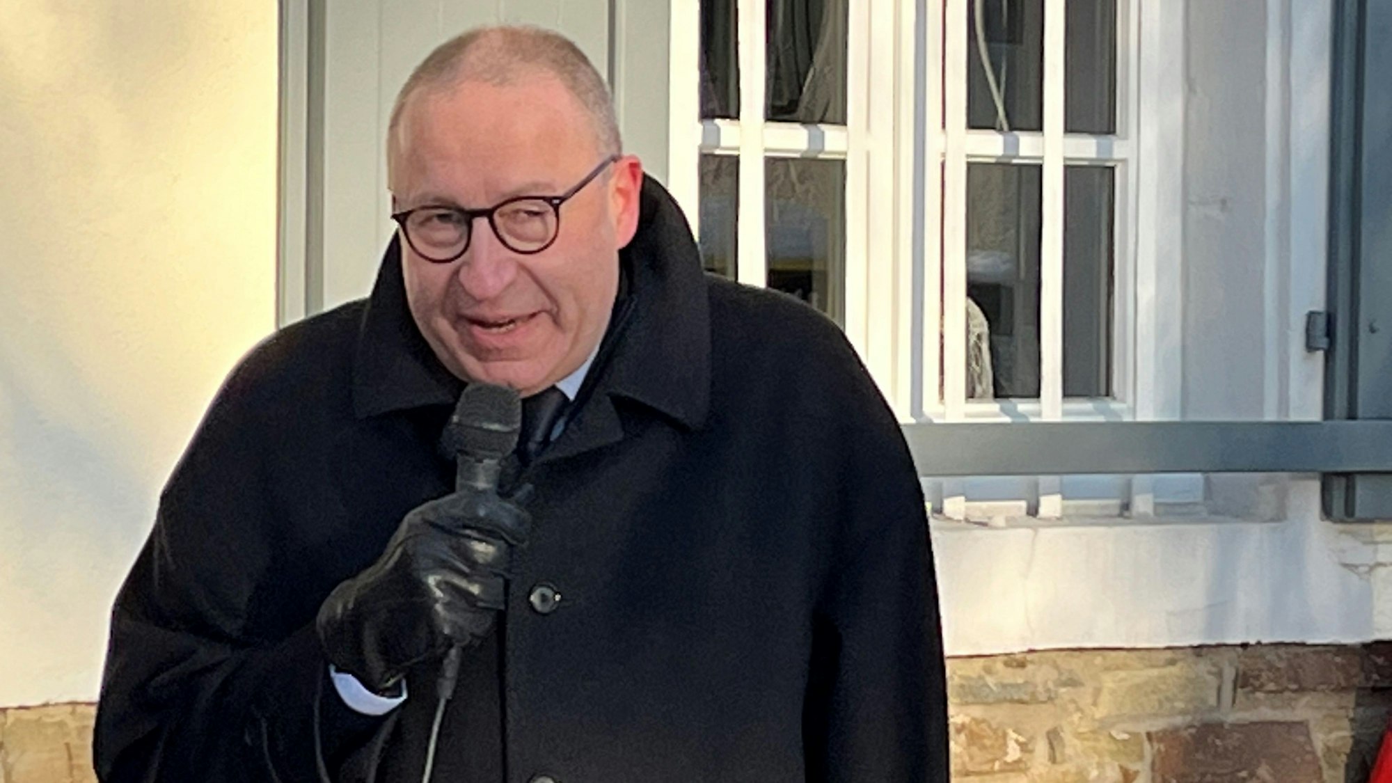 Ein Mann im schwarzen Mantel und Lederhandschuhen spricht in ein Mikrophon.