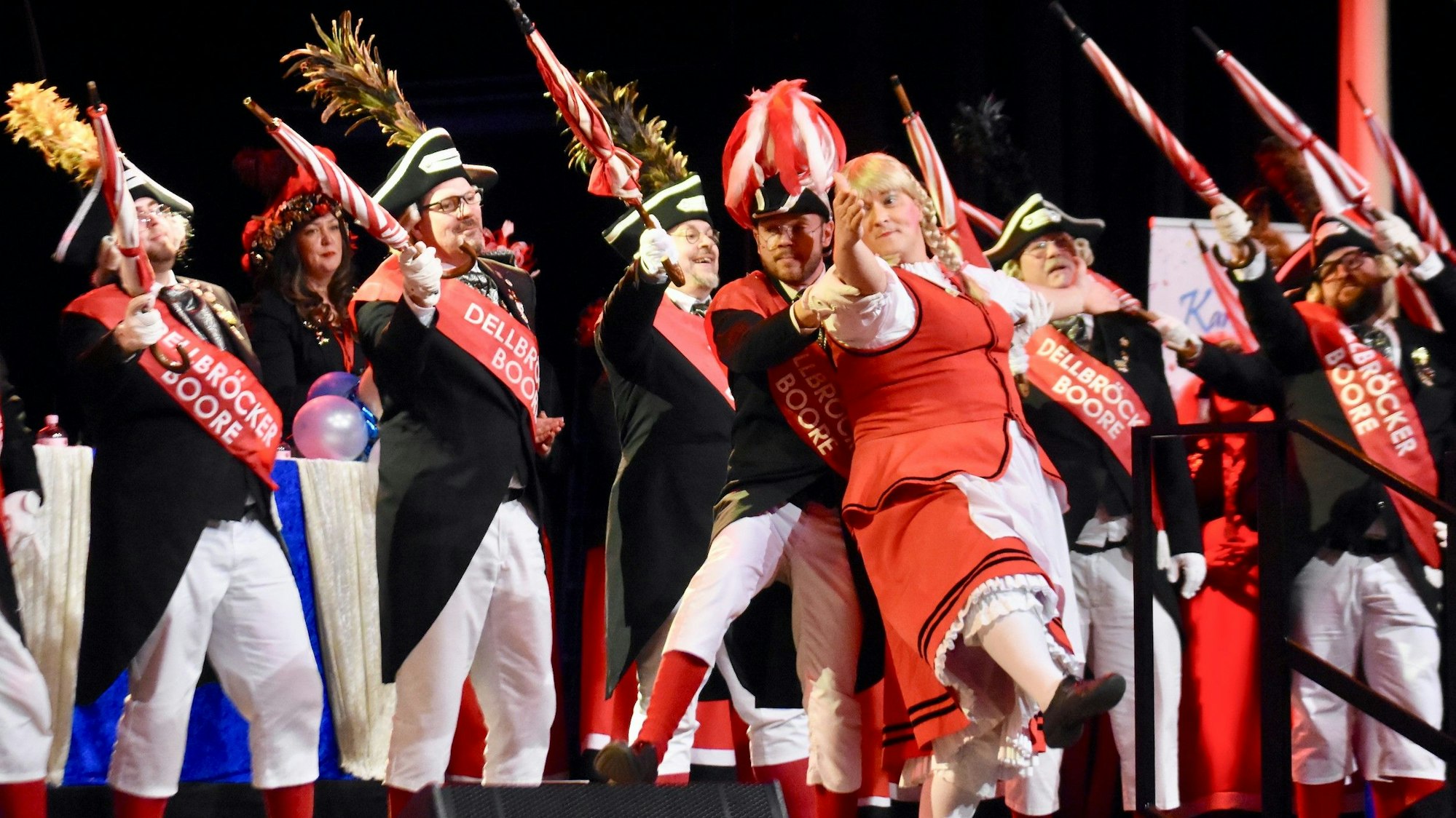 Ein Karnevalstanzkorps auf einer Bühne; im Vordergrundd tanzt ein Mann im Frauenkostüm mit blonder Zopfperücke.