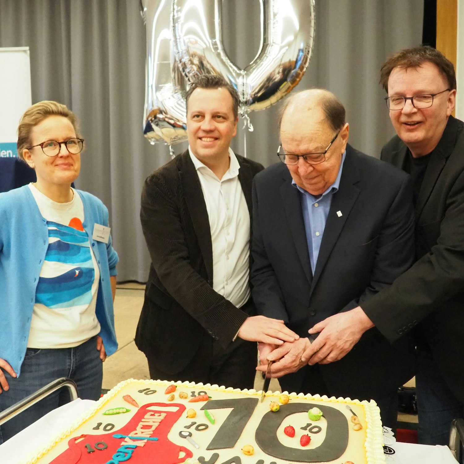 Die Genannten schneiden gemeinsam mit einem Messer in einen quadratischen Kuchen, auf dem die 10 in vielen Variationen zu sehen ist.