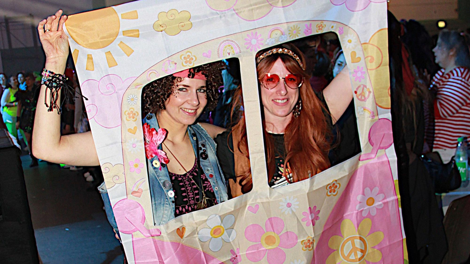 Zwei als Hippies verkleidete Frauen heben ein Tuch, das einen Bulli darstellen soll, und schauen aus den in Fensterform ausgeschnittenen Löchern.