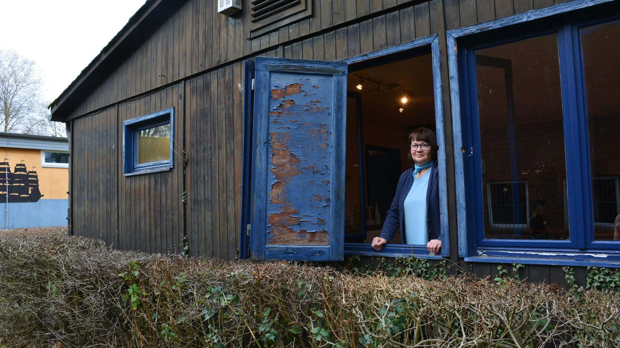 Eine Frau schaut aus einem Blockhaus. Am Fensterrahmen hängt ein lädierter Fensterladen.