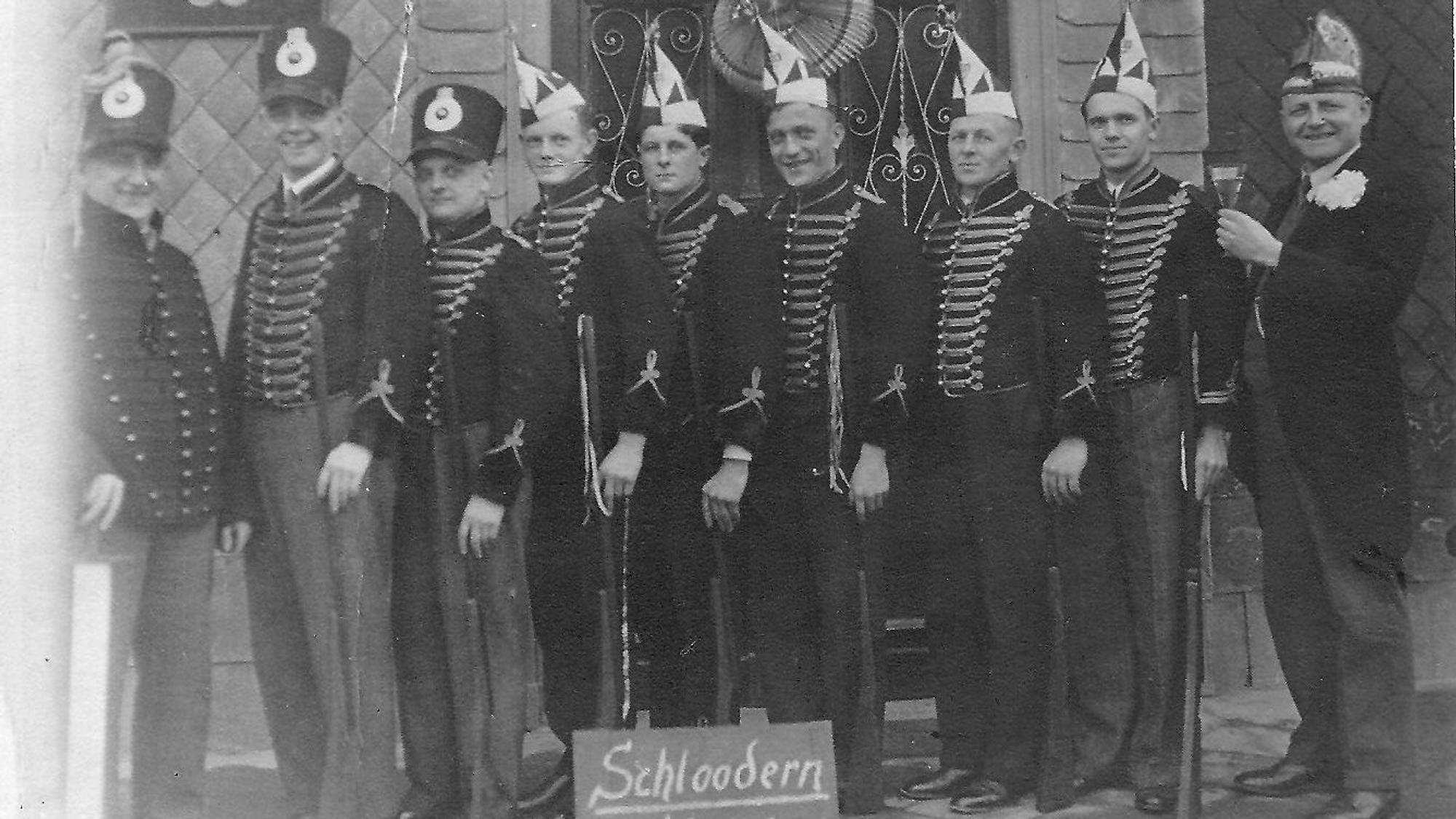 Eine historische Schwarz-Weiß-Aufnahme; Männer mit Karnevalsmützen stehen in einer Reihe.
