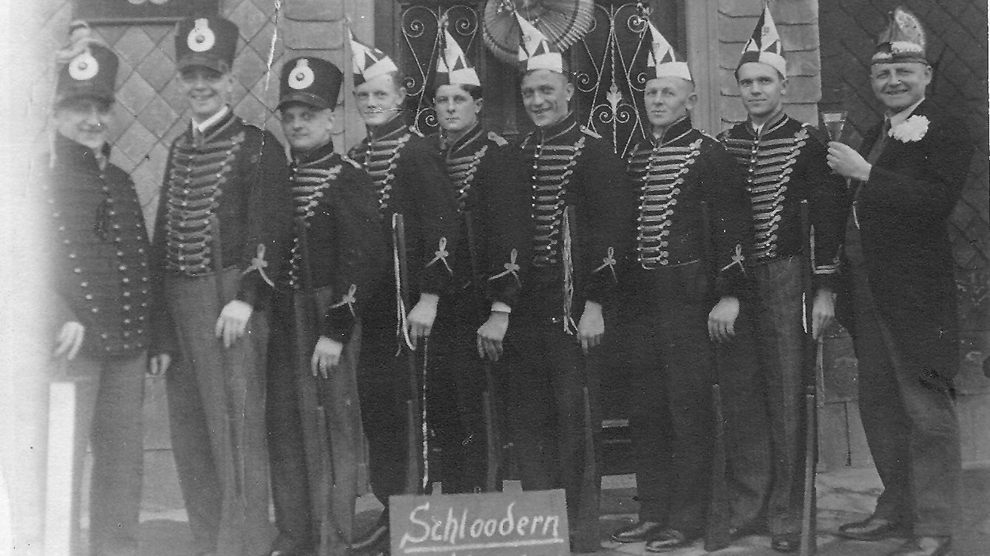 Eine historische Schwarz-Weiß-Aufnahme; Männer mit Karnevalsmützen stehen in einer Reihe.