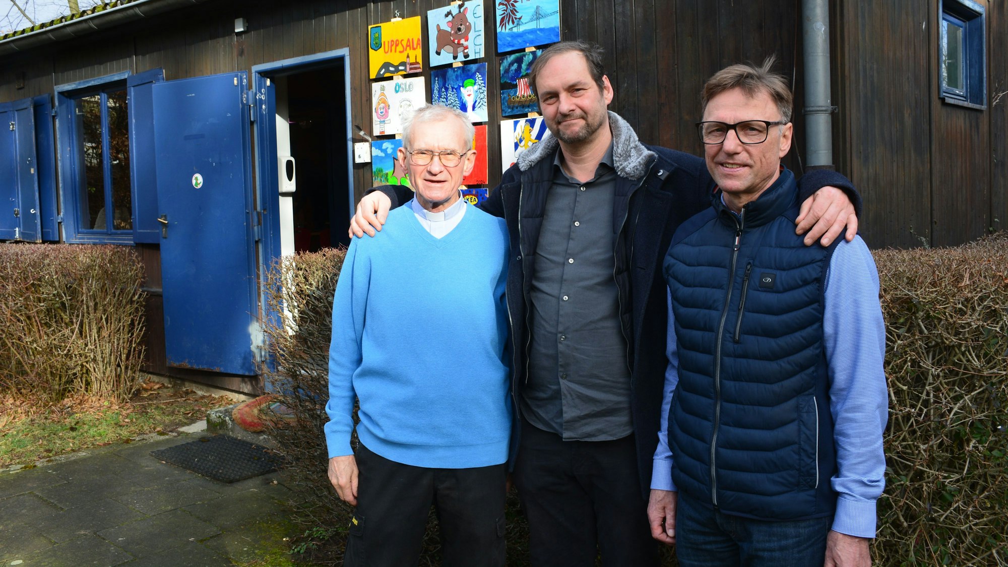 Drei Männer stehen vor einem braunen Blockhaus mit blauen Fensterläden und Türen.