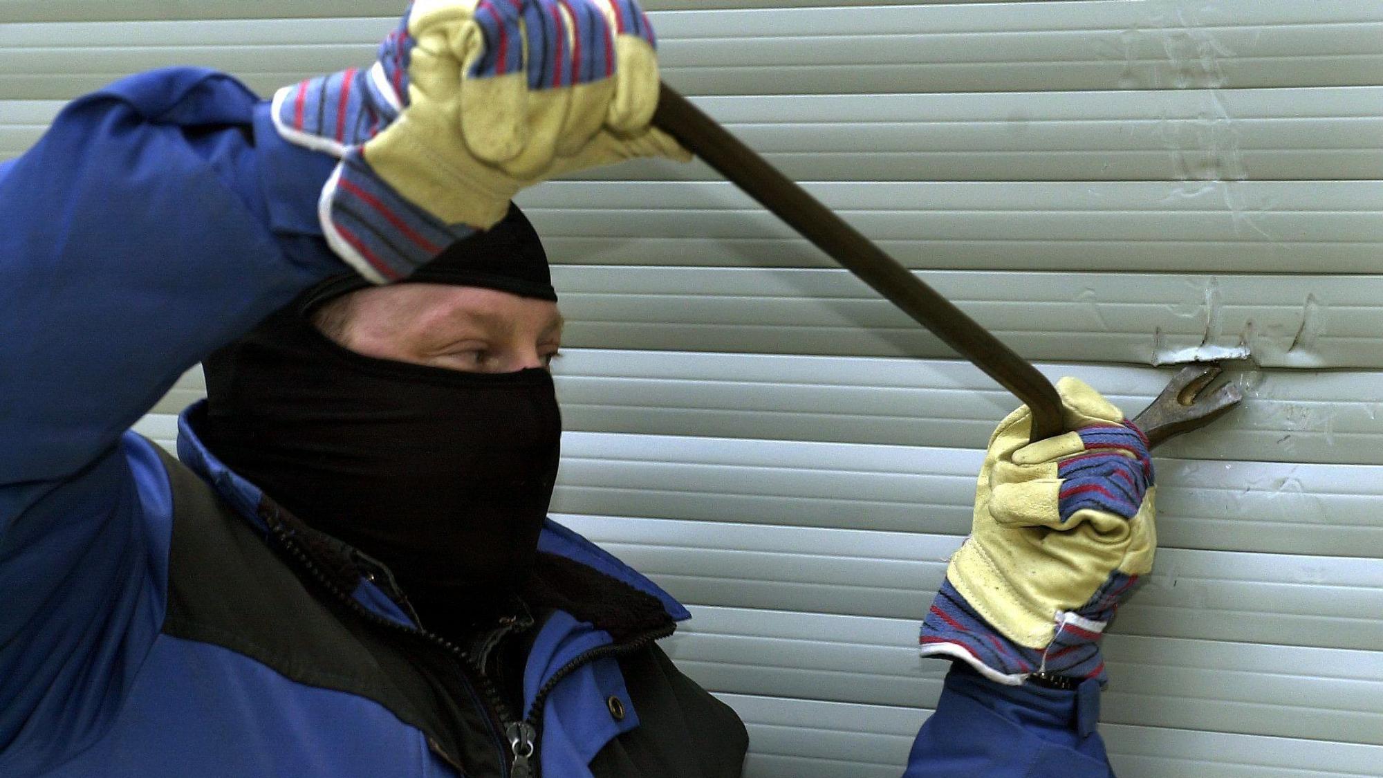 Ein mit Sturmmaske maskierter Mann setzt ein Brecheisen an einer geschlossenen Rollade an.