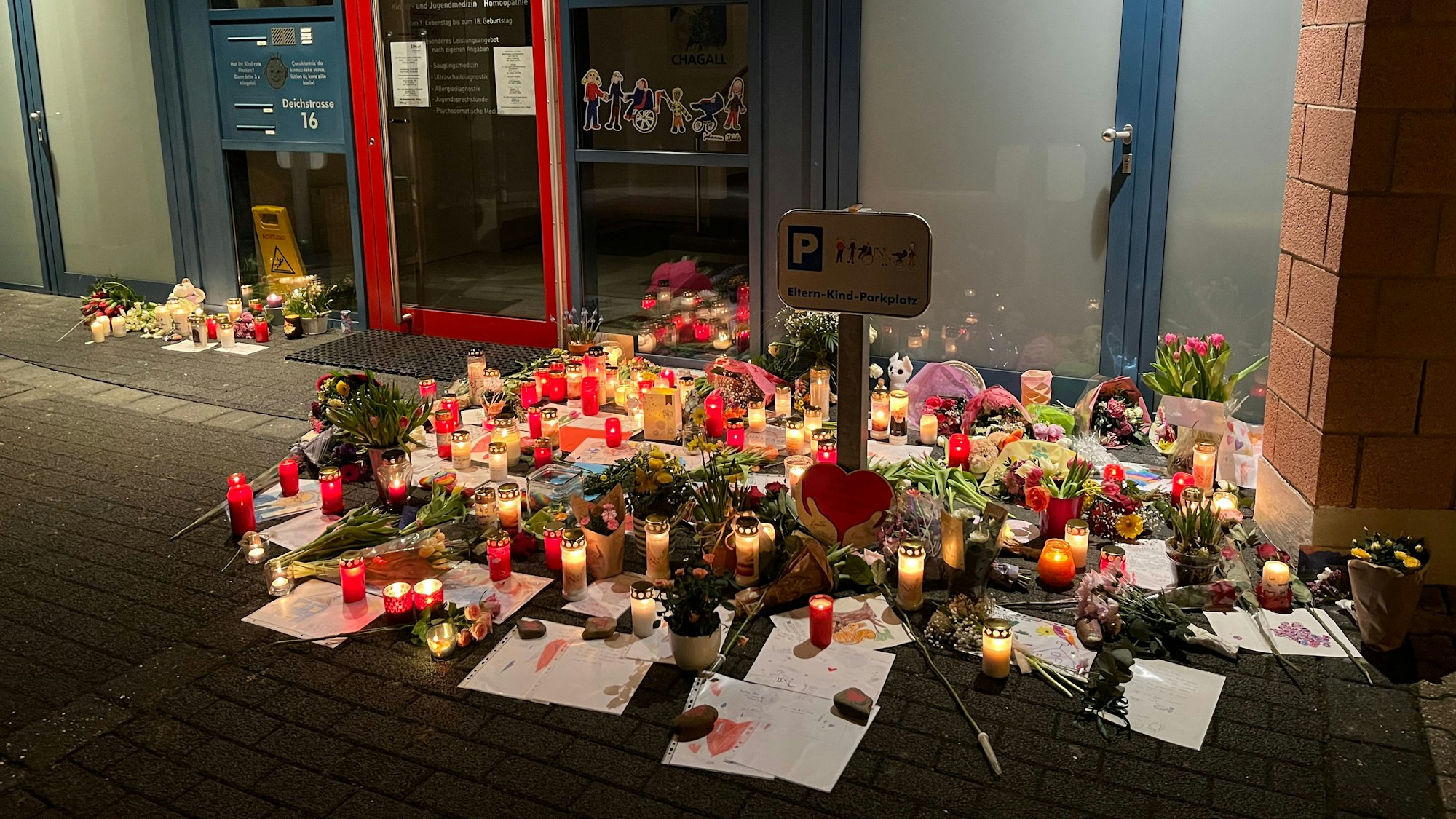 Kerzen stehen vor einer Tür zu einer Kinderarztpraxis, dazu selbst gemalte Bilder von Kindern.