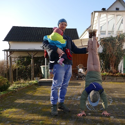 Ein Mann mit Mütze und Brille steht in einem Garten. Er hält ein Kind auf dem Arm, dessen Gesicht von der Kamera abgewandt ist. Ein weiteres Kind macht einen Handstand und schaut dabei in Richtung des Bodens.