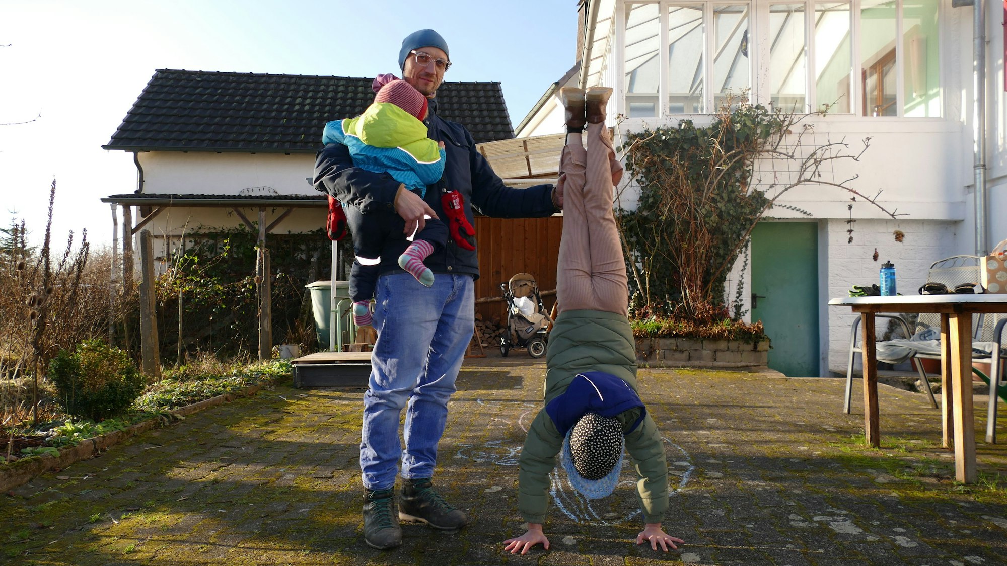 Ein Mann mit Mütze und Brille steht in einem Garten. Er hält ein Kind auf dem Arm, dessen Gesicht von der Kamera abgewandt ist. Ein weiteres Kind macht einen Handstand und schaut dabei in Richtung des Bodens.