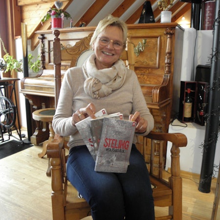 Ute Mainz sitzt in einem Stuhl und hält ihr neues Buch in Händen, im Hintergrund ist ein Klavier zu sehen.