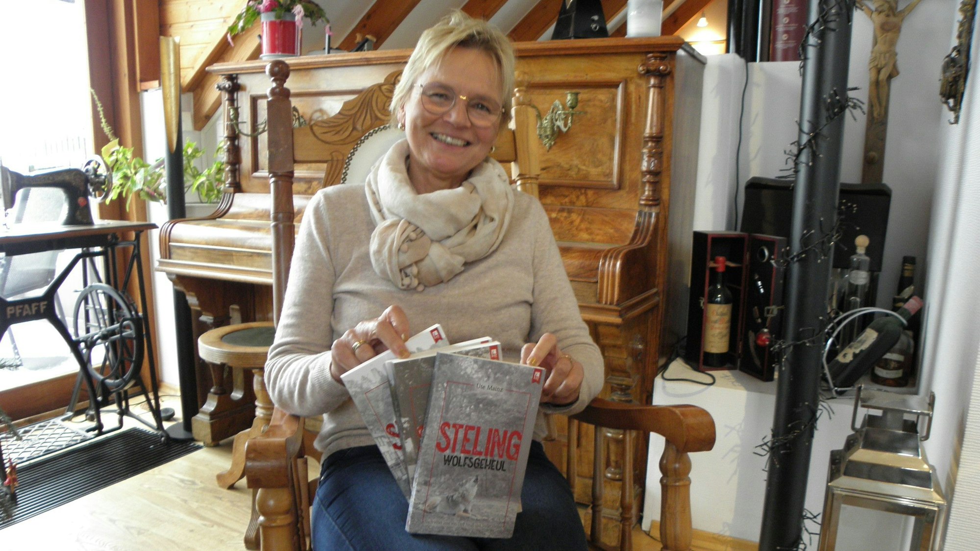 Ute Mainz sitzt in einem Stuhl und hält ihr neues Buch in Händen, im Hintergrund ist ein Klavier zu sehen.