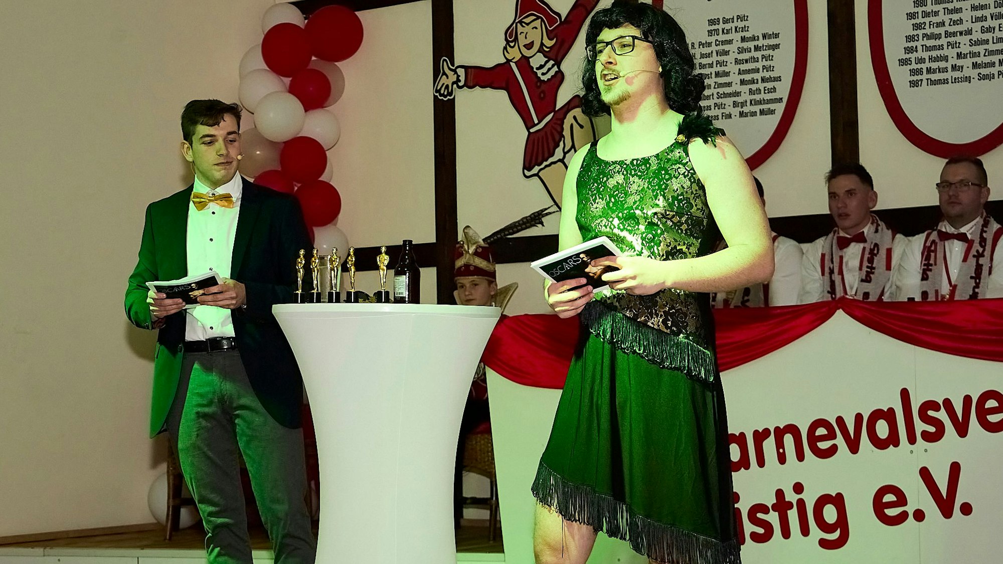 Ein Mann im Frack und ein Mann in festlicher Frauenkleidung stehen an einem Tisch, auf dem goldene Oscar-Figuren stehen.