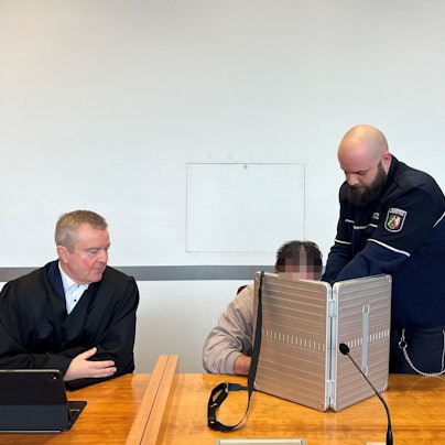 Der Angeklagte wird von einem Justizbediensteten in den Sitzungssaal des Bonner Landgerichts gebracht. Er verbirgt sein Gesicht hinter einem silberfarbenen schmalen Koffer, neben ihm sitzt sein Anwalt in schwarzer Robe.