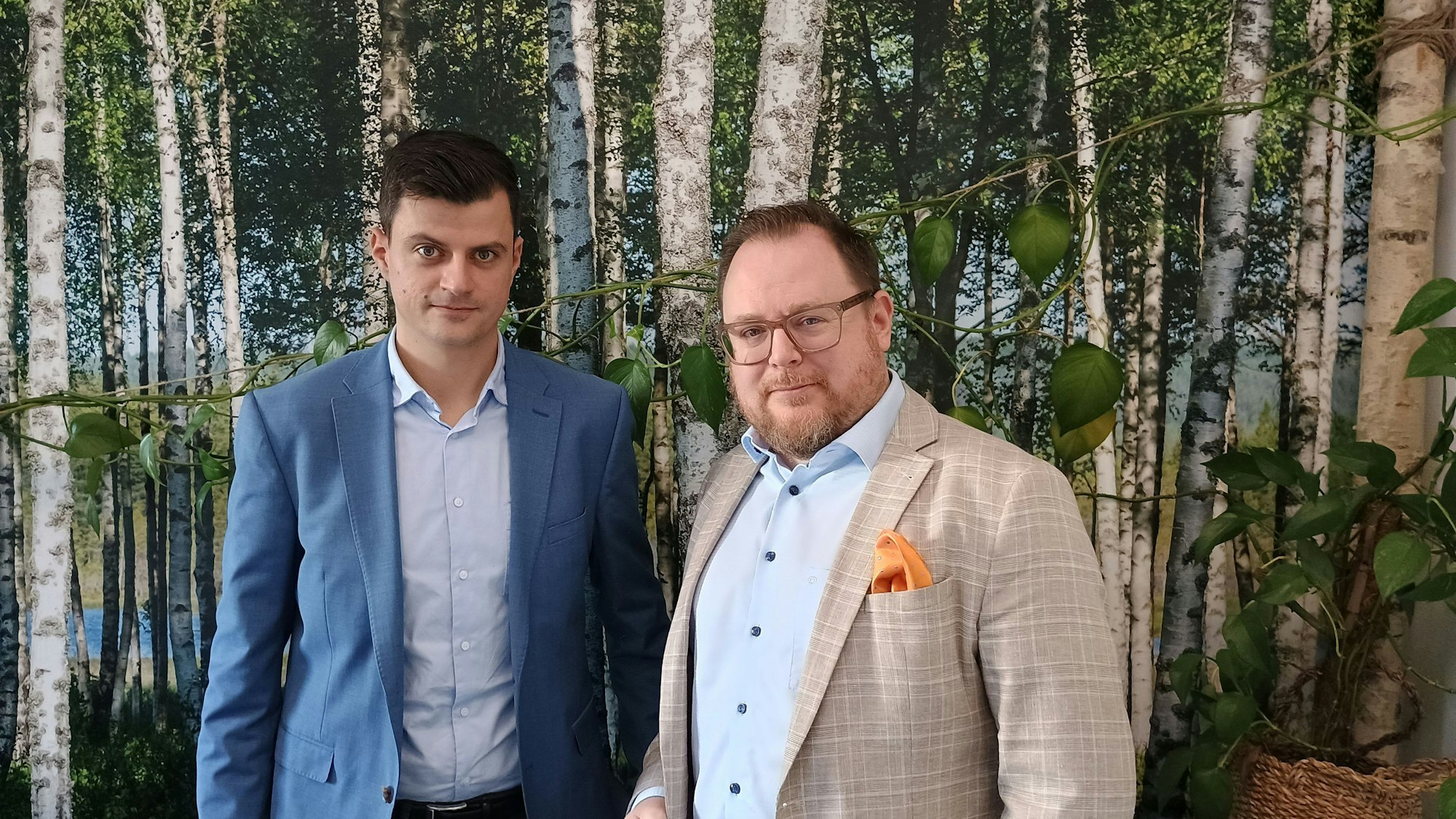 Die beiden Männer tragen Anzüge. Sie stehen vor einer Tapete, auf denen Bäume abgebildet sind.