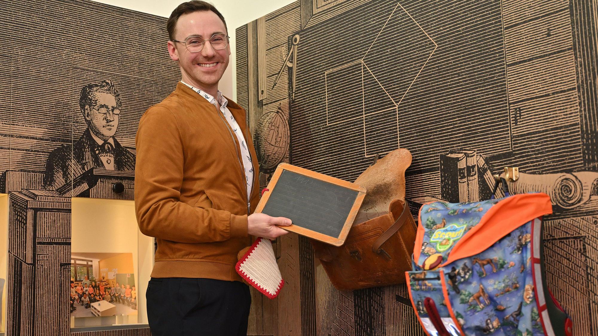 Ein junger Mann hat eine alte Schiefertafel in der Hand. An der Wand hängen ein alter und ein moderner Tornister.