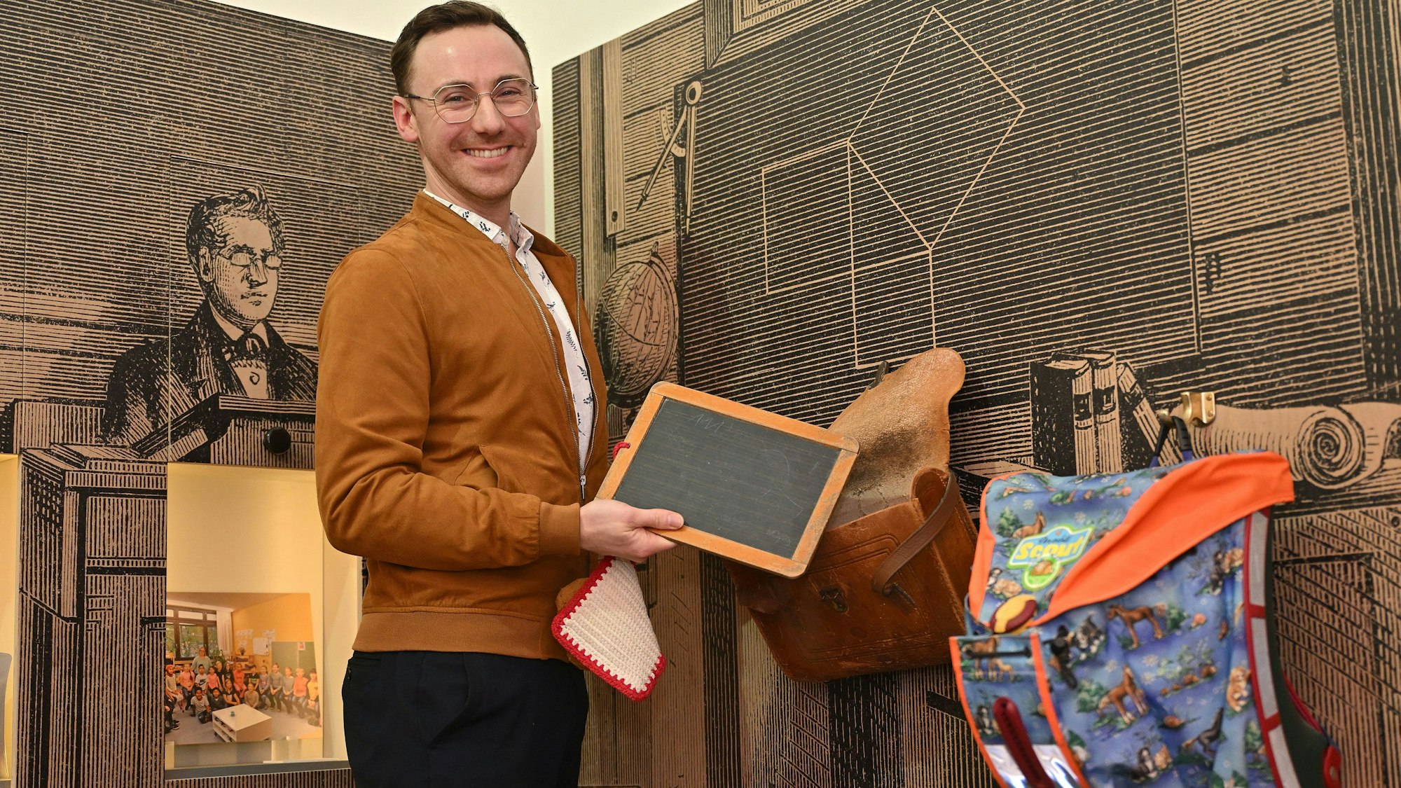 Ein junger Mann hat eine alte Schiefertafel in der Hand. An der Wand hängen ein alter und ein moderner Tornister.