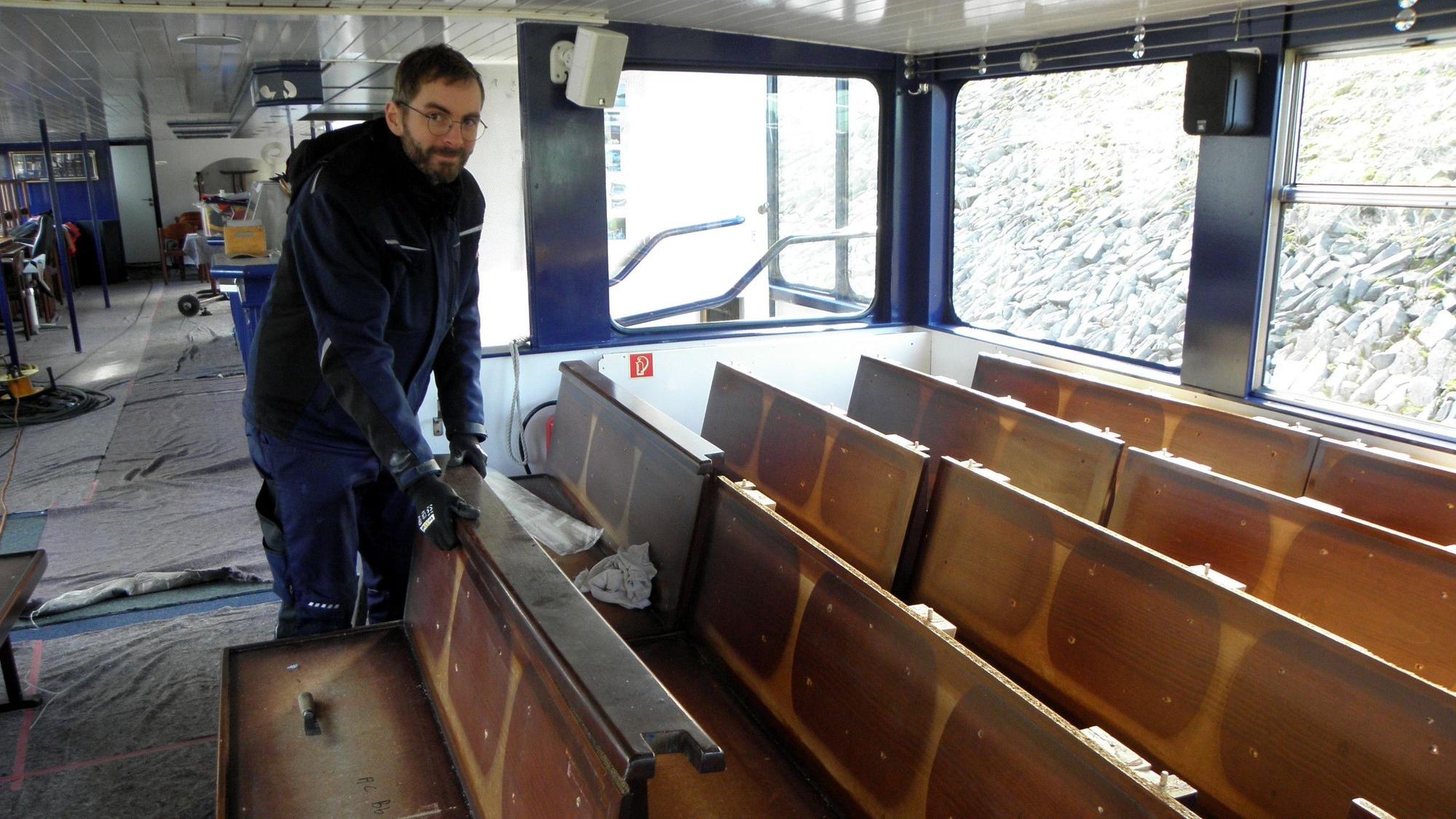 Ein Mann steht im Innenraum des Rursee-Schiffes Aachen an Sitzreihen aus Holz. Die Polster sind entfernt, sie werden durch neue ersetzt.
