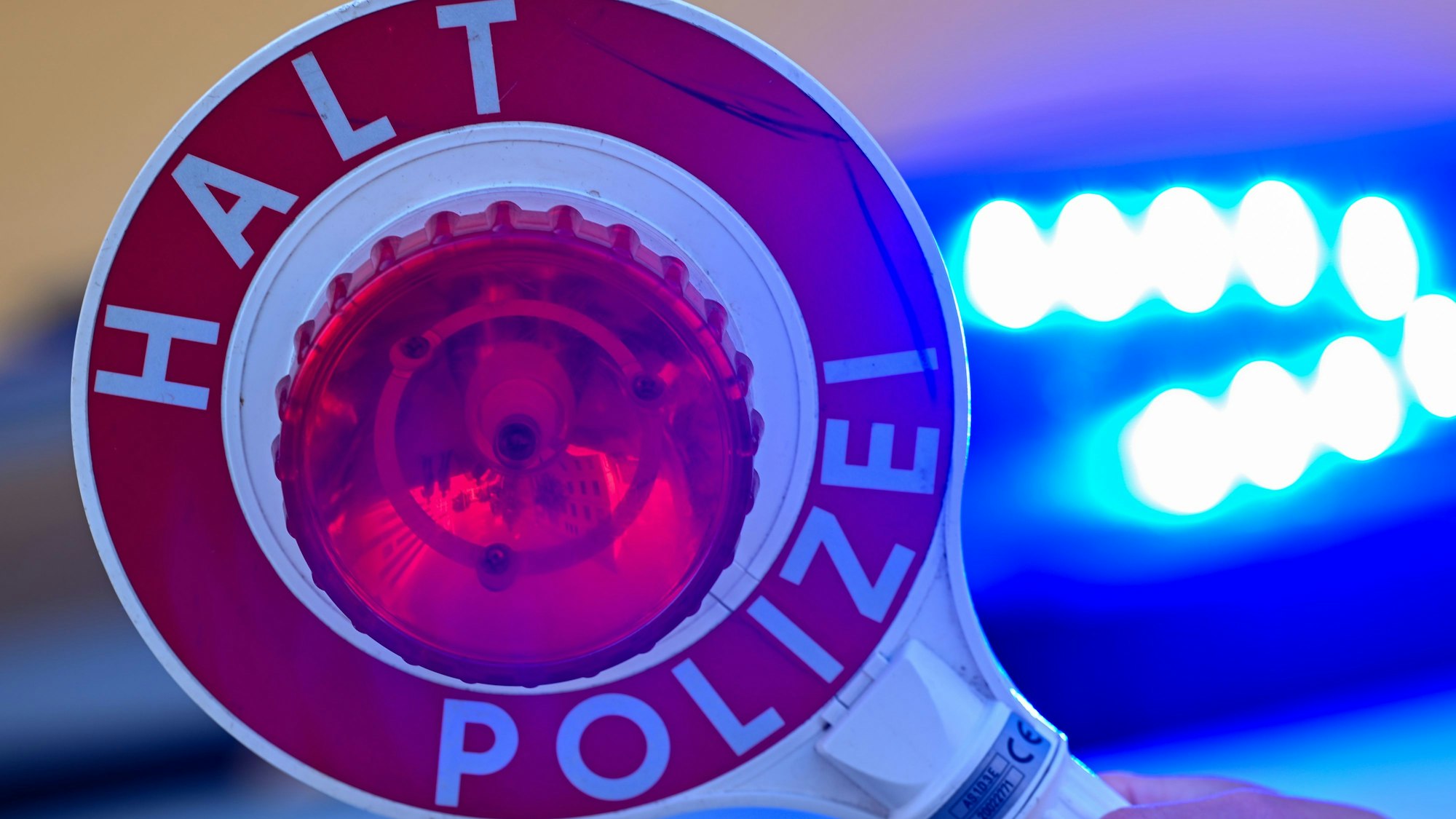 Das Symbolbild zeigt eine Kelle der Polizei mit der Aufschrift „Halt Polizei“ vor dem Blaulicht eines Streifenwagens.