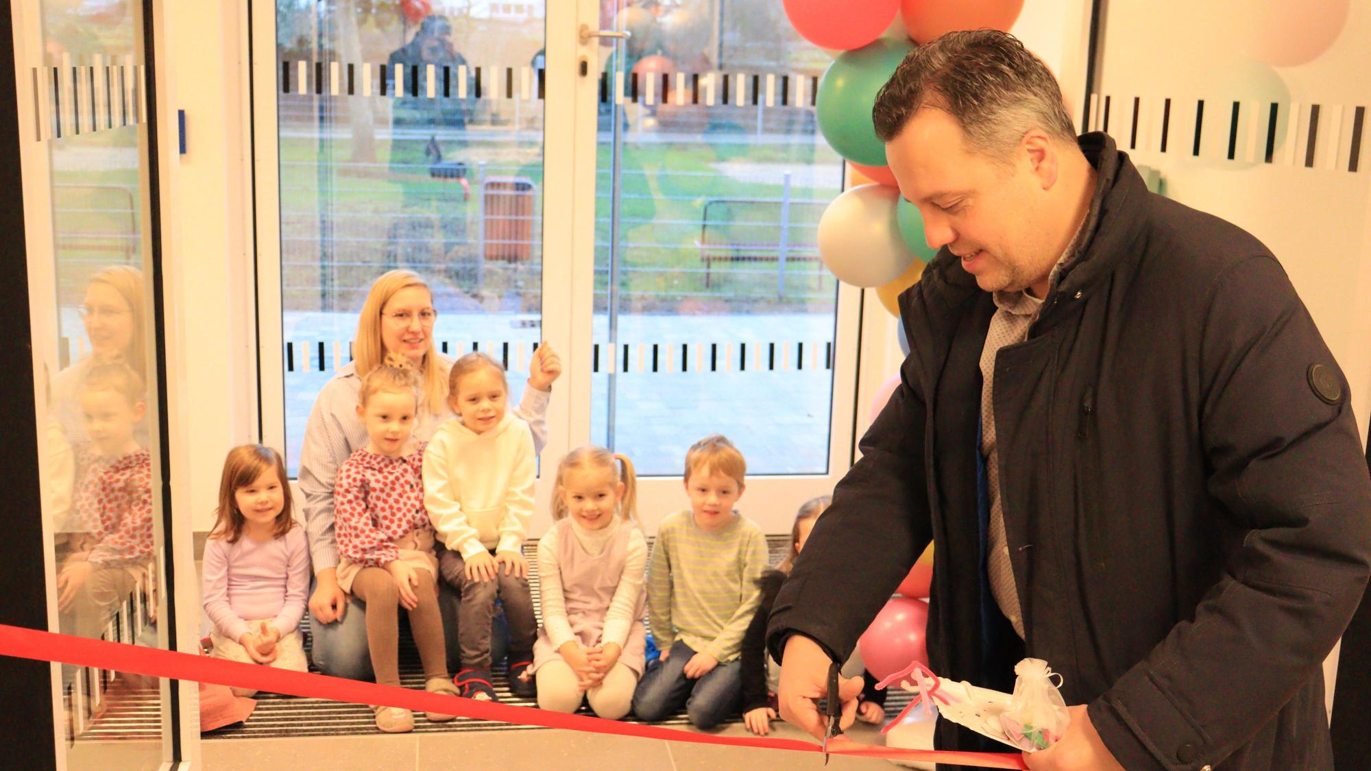 Sacha Reichelt schneidet ein rotes Band durch. Mehrere Kinder und eine Erzieherin im Hintergrund schauen zu.