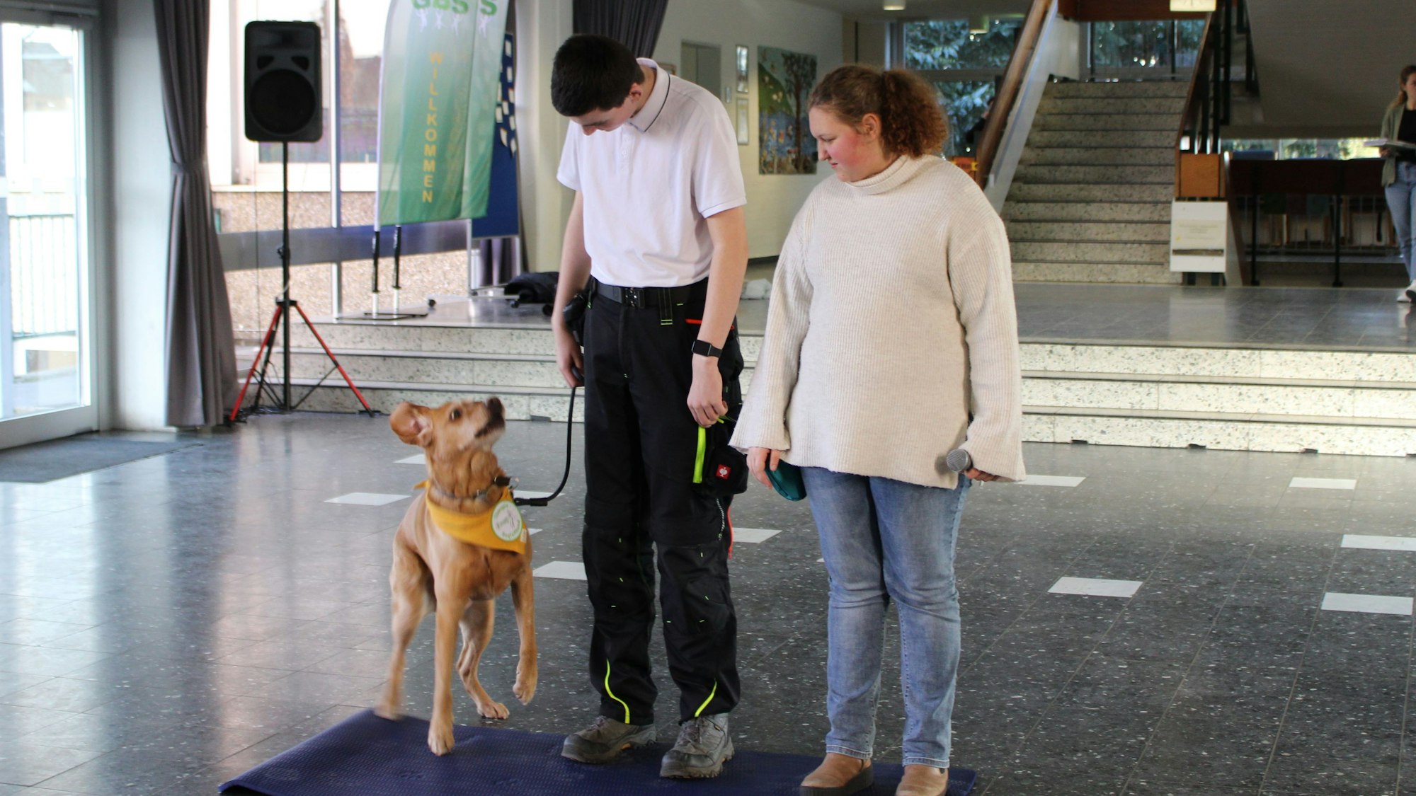 Eine Schülerin und ein Schüler stehen nebeneinander auf einer Matte im Eingangsbereich einer Schule. Das Mädchen hält ein Mikrophon, der Junge einen Hund an der Leine, der gerade hochspringt.