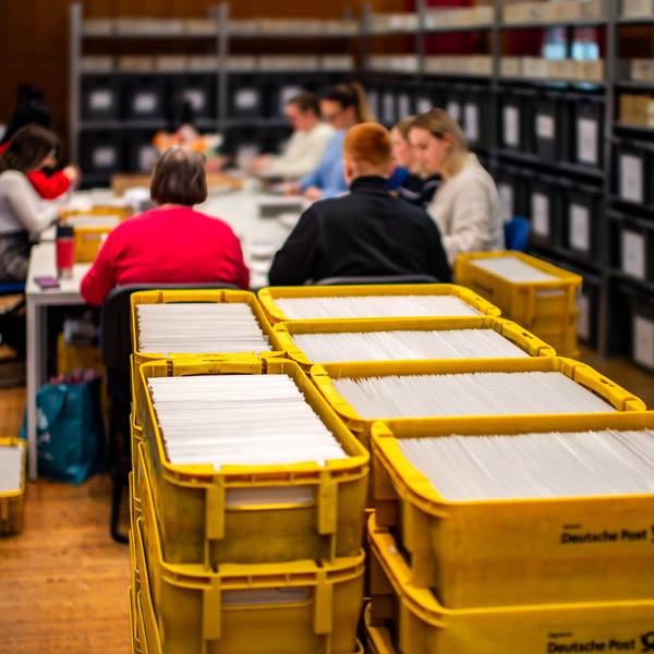Das Bild zeigt zahlreiche gelbe Postkisten im Vordergrund, die randvoll mit Wahlunterlagen sind.