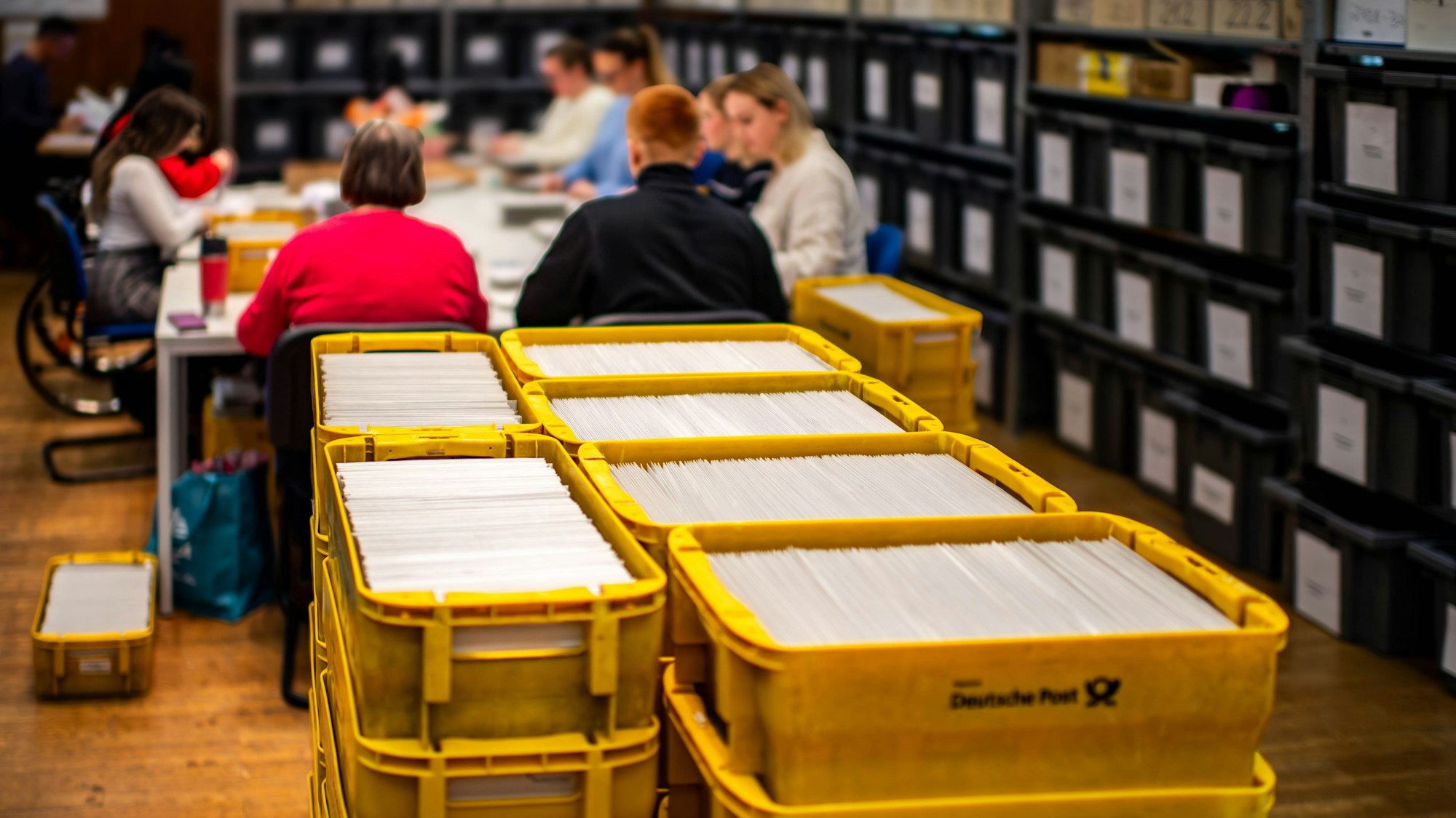 Das Bild zeigt zahlreiche gelbe Postkisten im Vordergrund, die randvoll mit Wahlunterlagen sind.