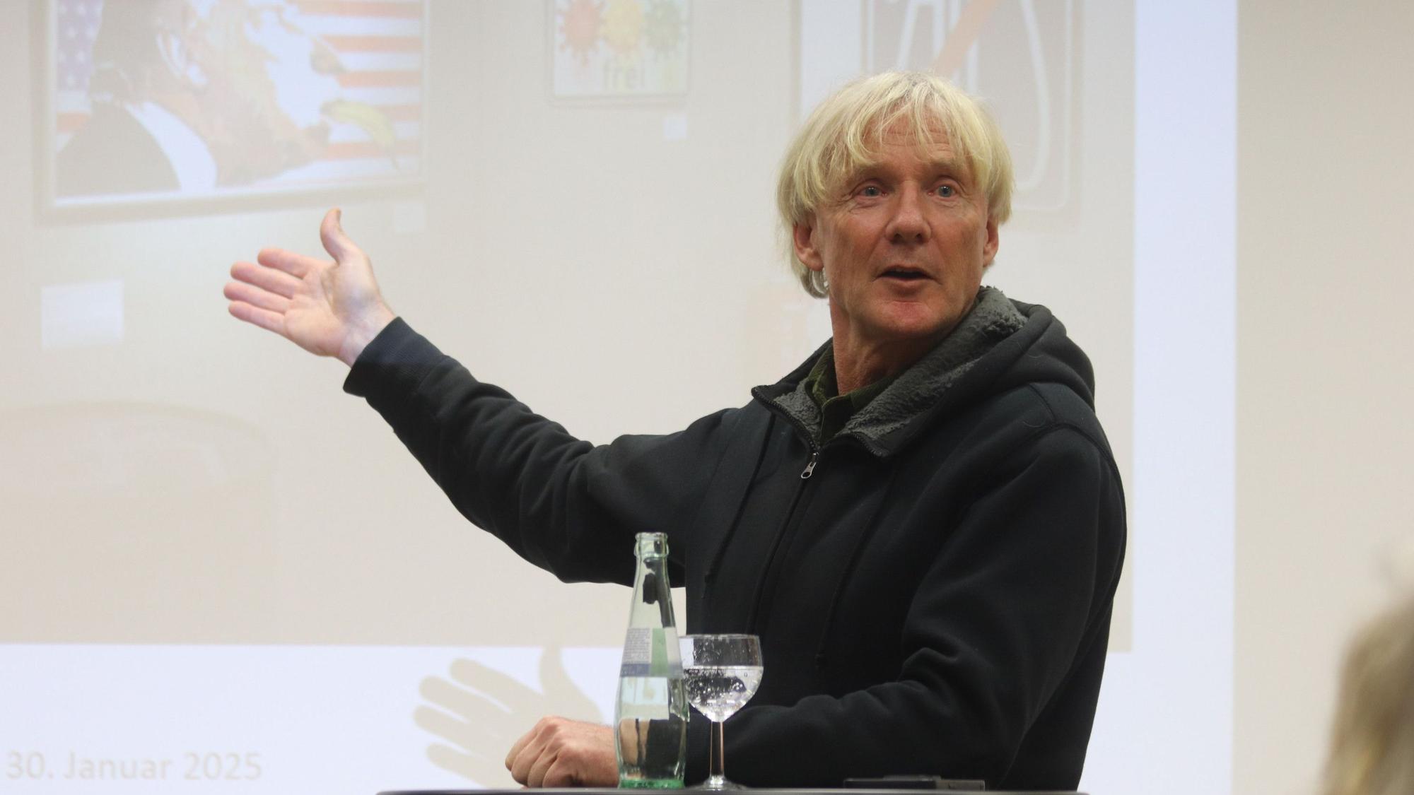 Der als Bananensprayer bekannte Thomas Baumgärtel steht gestikulierend vor einem an die Wand gebeamten Bild.