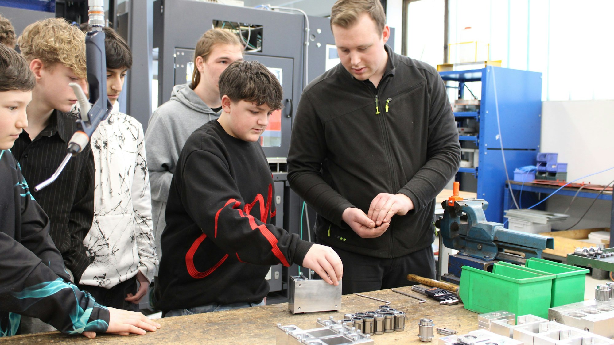 Ein Junge schraubt an einer Metallbox, ein Mitarbeiter leitet ihn an, seine Mitschüler schauen ihm zu.