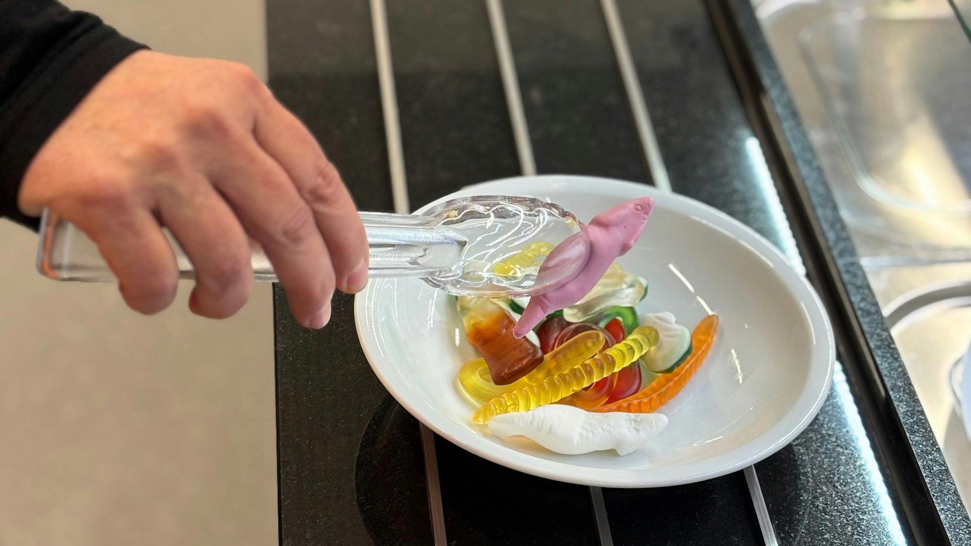 Auf einer Nahaufnahme hält eine Hand eine durchsichtige Greifzange, die Mäusespeck in Form einer Maus aus einer Schale mit Süßigkeiten gepackt hat.