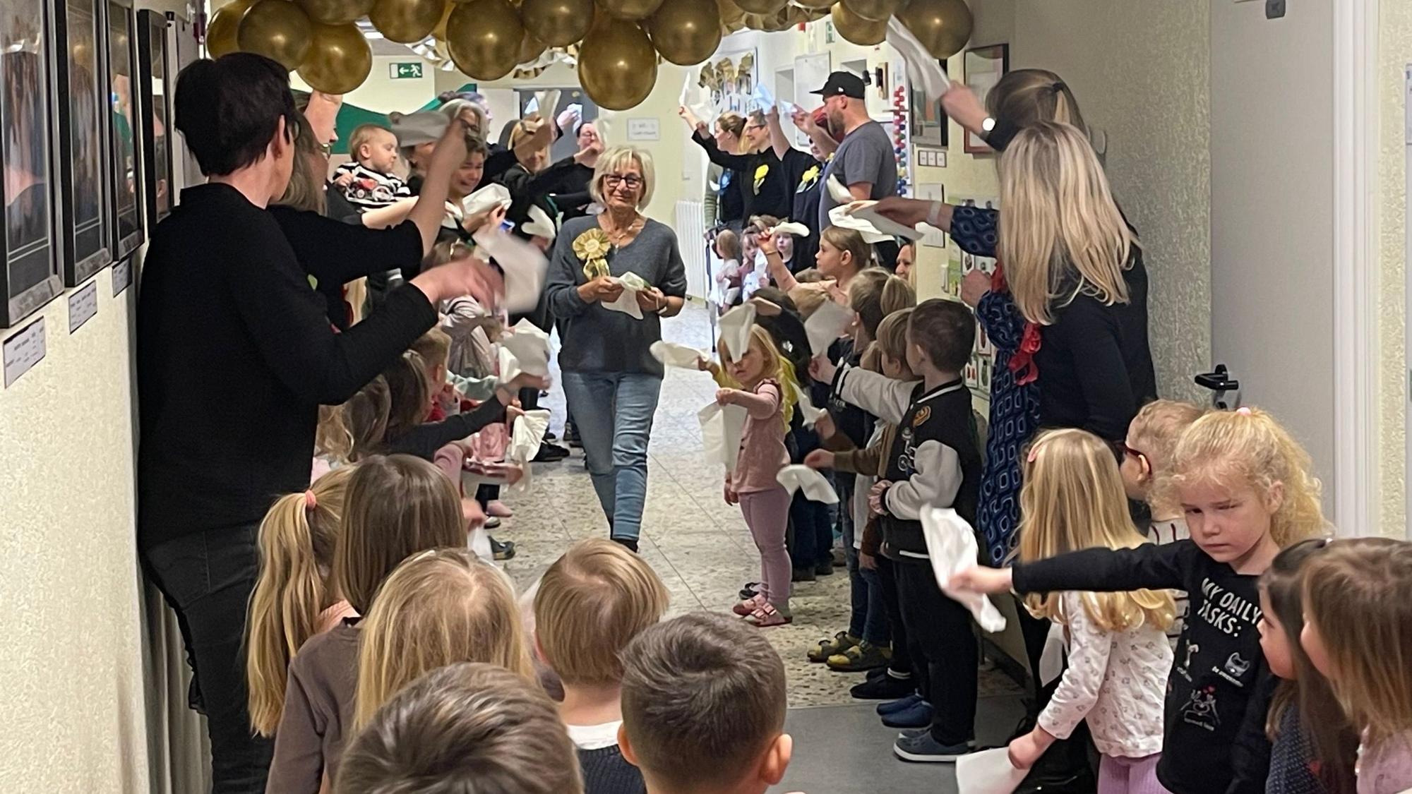 Zu sehen ist eine Frau, die durch ein Spalier von Kindern und Erwachsenen geht, die mit Taschentüchern winken.