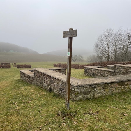 Eine Mauer steht auf einer Wiese, dahinter sind kahle Büsche und Hügel, die im Nebel verschwinden zu sehen.