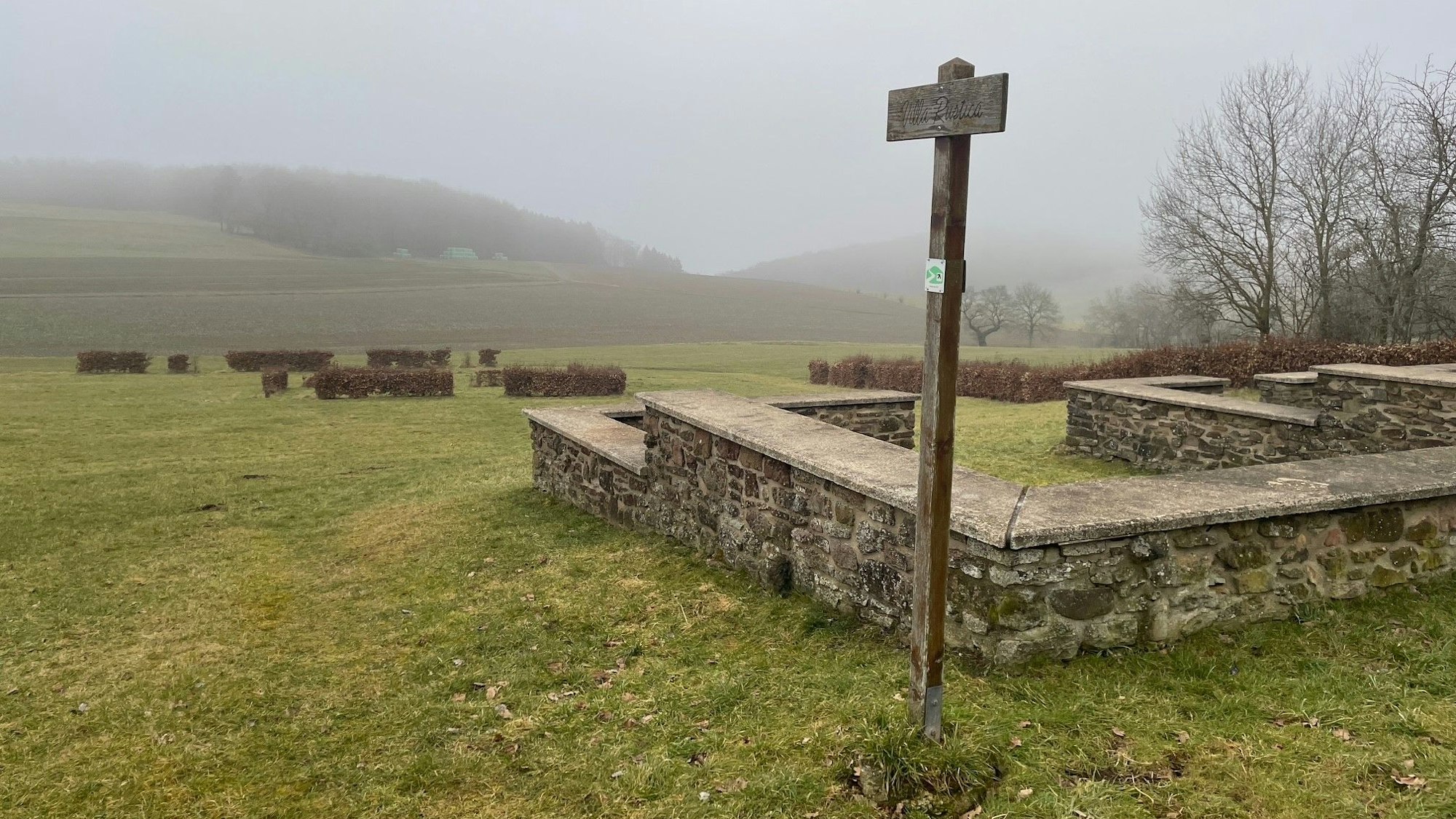 Eine Mauer steht auf einer Wiese, dahinter sind kahle Büsche und Hügel, die im Nebel verschwinden zu sehen.