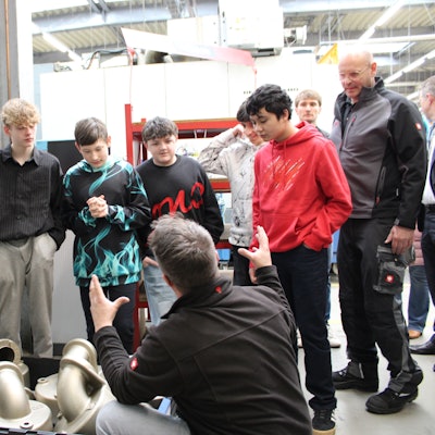 Sieben Jungen stehen in einer Fabrikhalle, ein Mitarbeiter hockt gestikulierend vor ihnen.