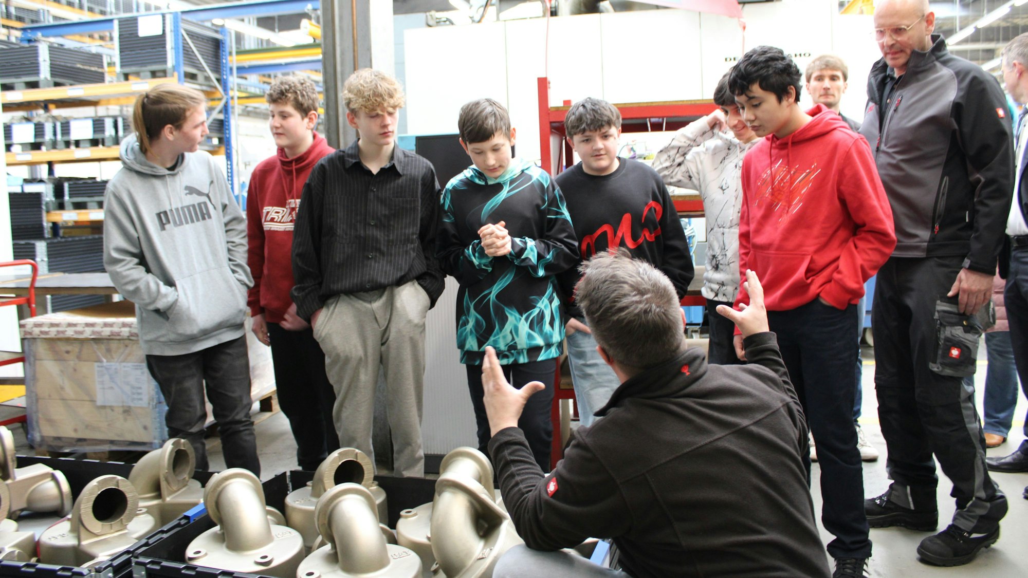 Sieben Jungen stehen in einer Fabrikhalle, ein Mitarbeiter hockt gestikulierend vor ihnen.
