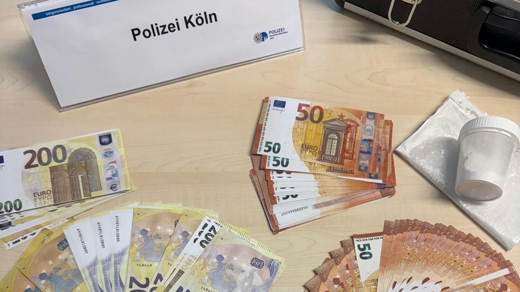 Das Foto zeigt falsche Geldscheine, eine Dose mit Betäubungsmitteln und ein Schild, auf dem „Polizei Köln“ steht.