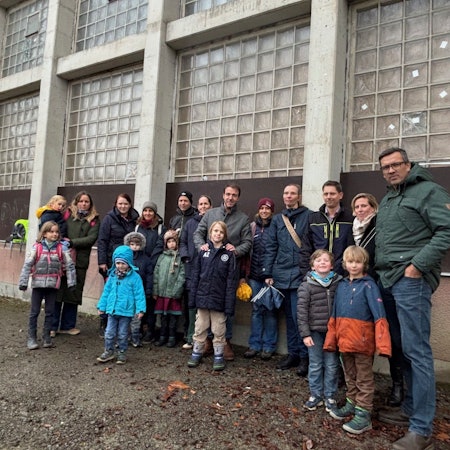 Auf dem Bild ist eine Gruppe von Eltern mit ihrem Kindern vor einer alten Sporthalle zu sehen.