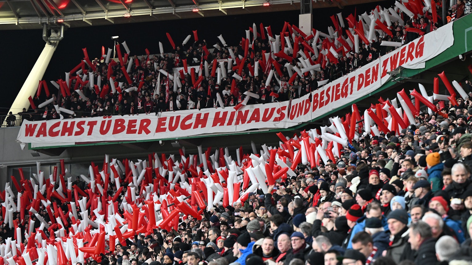 Im Kölner Fanblock hängt ein Spruchband mit der Aufschrift „Wachst über Euch hinaus und besiegt den Zwerg“.