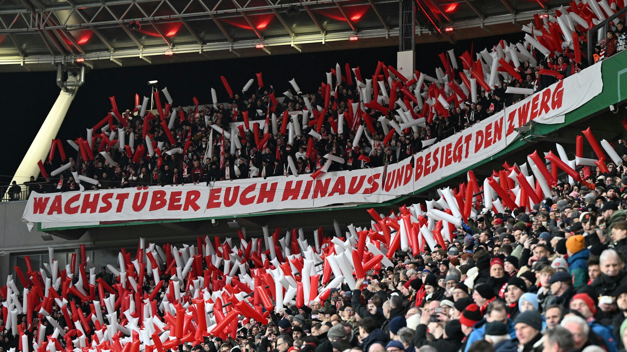 Im Kölner Fanblock hängt ein Spruchband mit der Aufschrift „Wachst über Euch hinaus und besiegt den Zwerg“.
