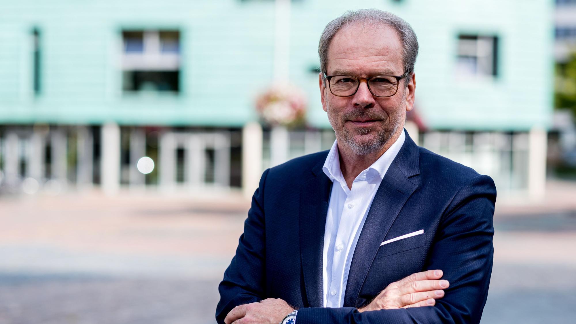 Der Bürgermeister trägt einen dunkelblauen Anzug und hat die Arme verschränkt. Im Hintergrund verschwommen die Stadtbibliothek.