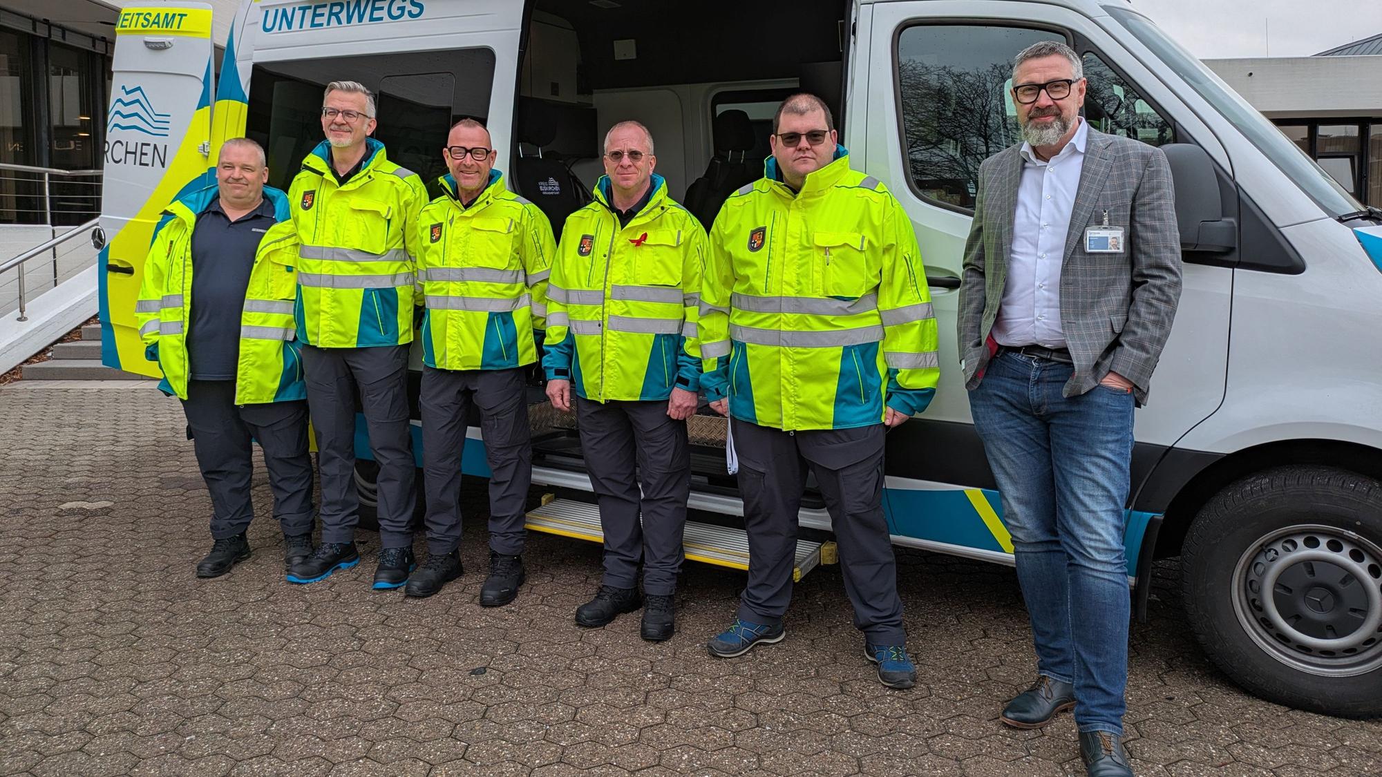 Eine Gruppe Männer in neongelben Jacken steht vor einem Fahrzeug, daneben steht ein Mann in Sakko, Hemd und Jeans.