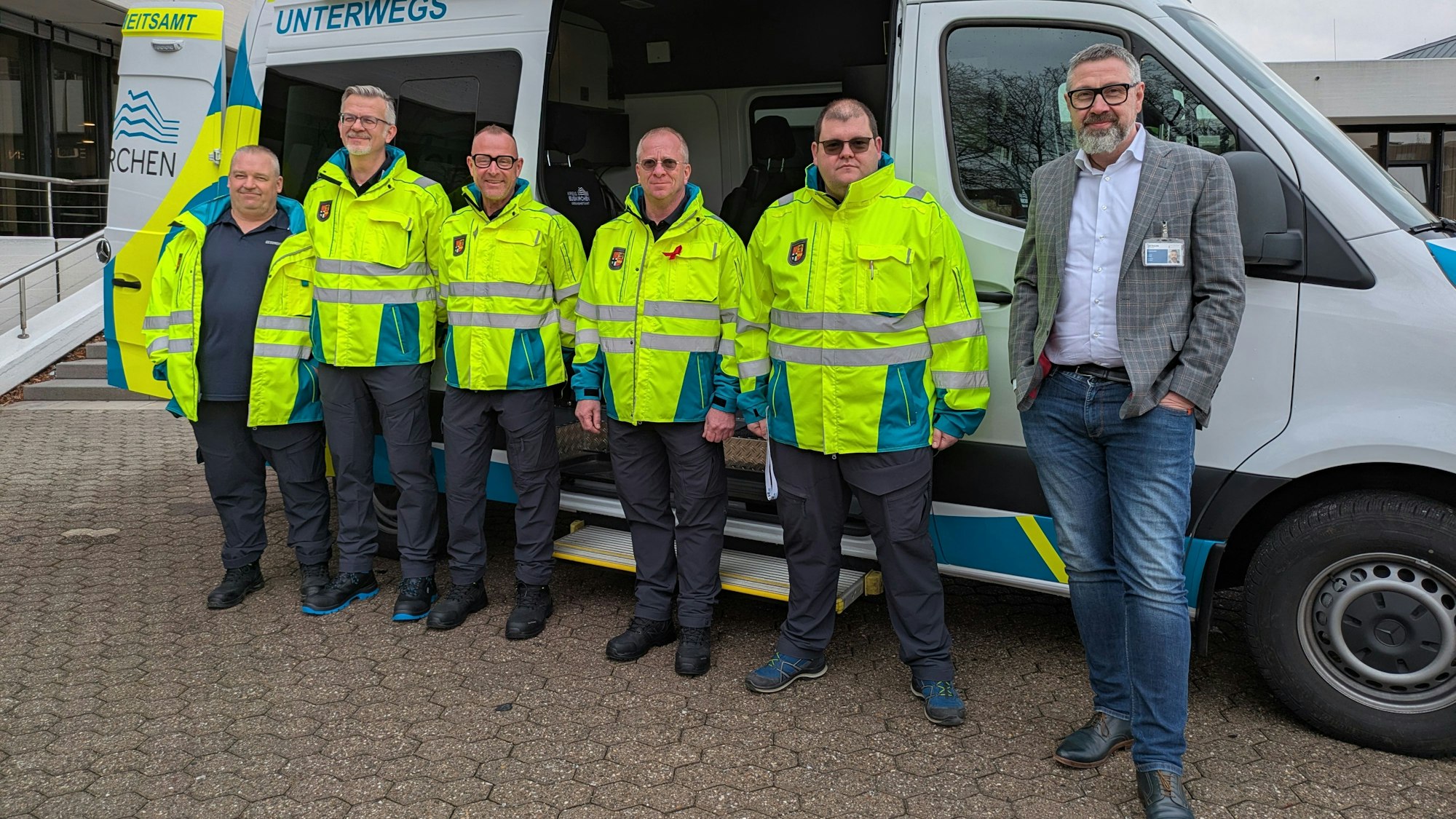 Eine Gruppe Männer in neongelben Jacken steht vor einem Fahrzeug, daneben steht ein Mann in Sakko, Hemd und Jeans.