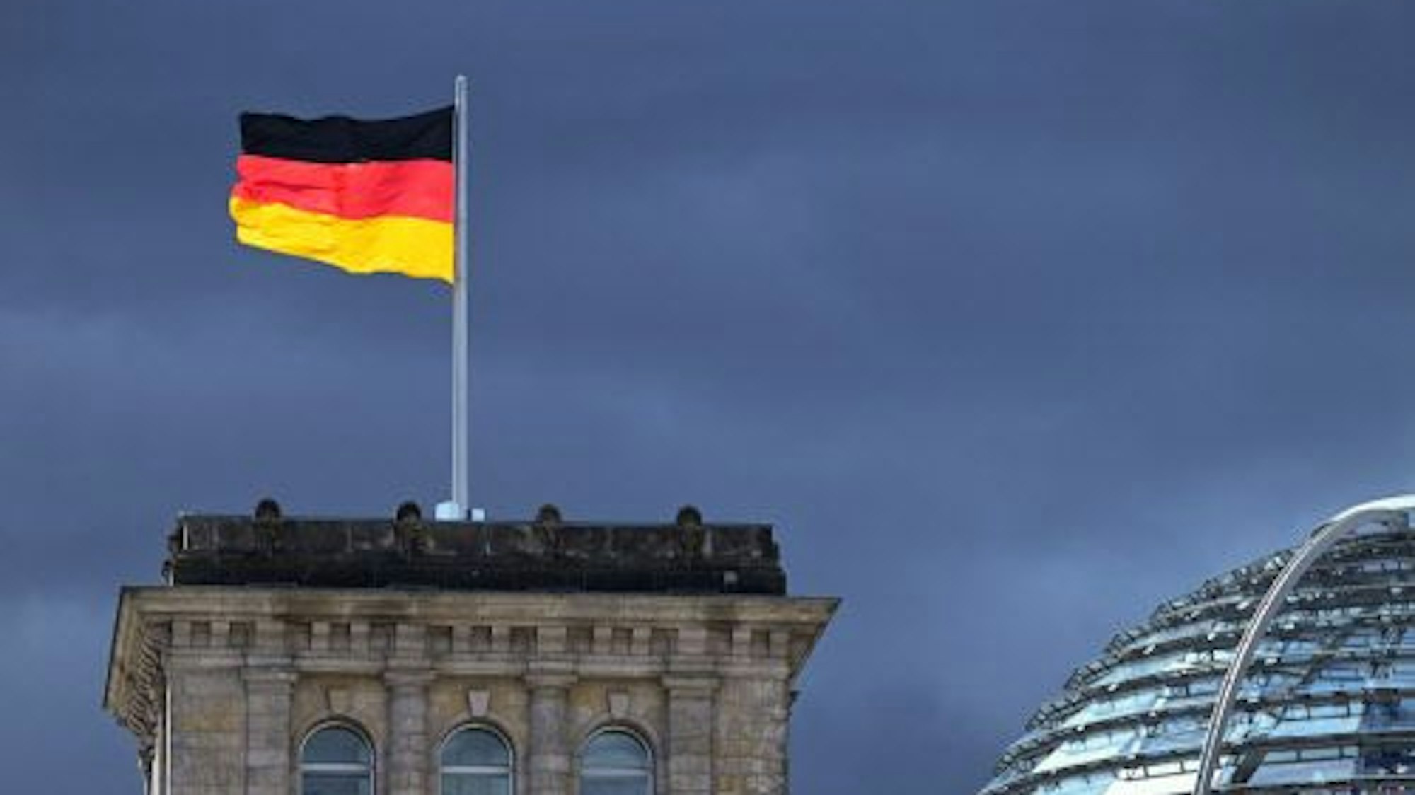 Eine der Flaggen auf dem Reichstag leuchtet bei starkem Wind wehend vor der Kulisse von dunklen Regenwolken. (Symbolbild)